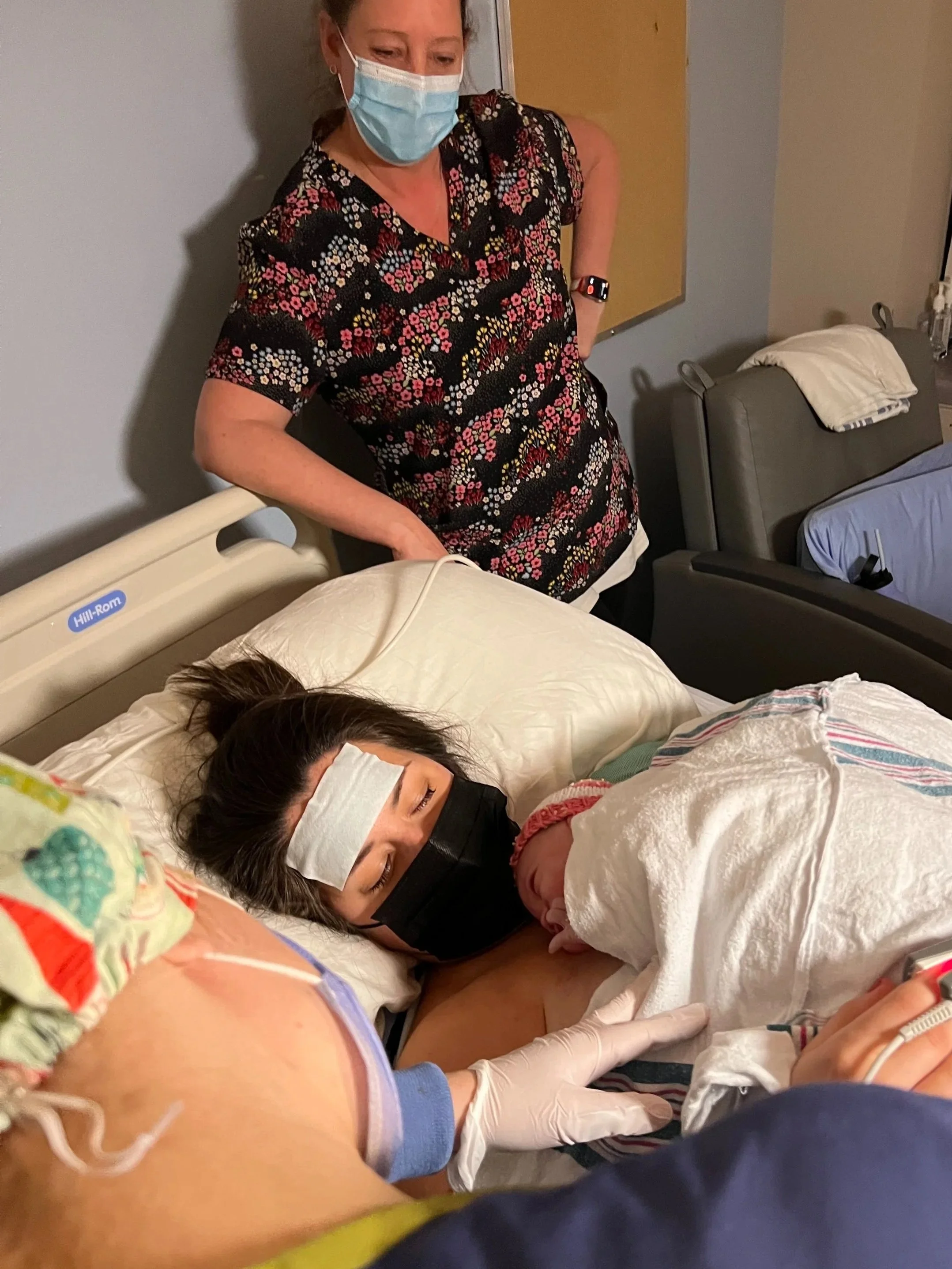 A woman in a floral shirt and wearing a face mask stands beside a hospital bed holding a newborn baby wrapped in a blanket, with a woman lying in the bed wearing a face mask and a hospital gown.