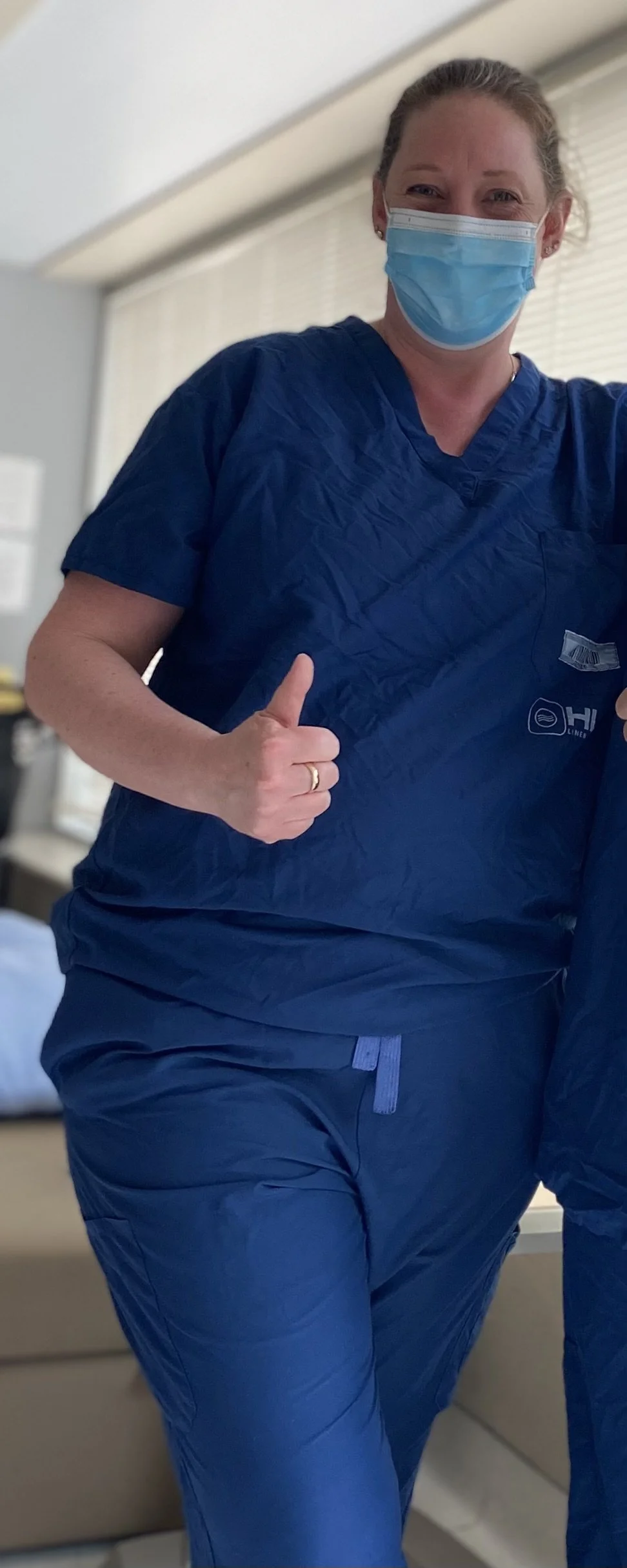 A woman in blue medical scrubs and a face mask shows a thumbs-up gesture inside a medical facility.