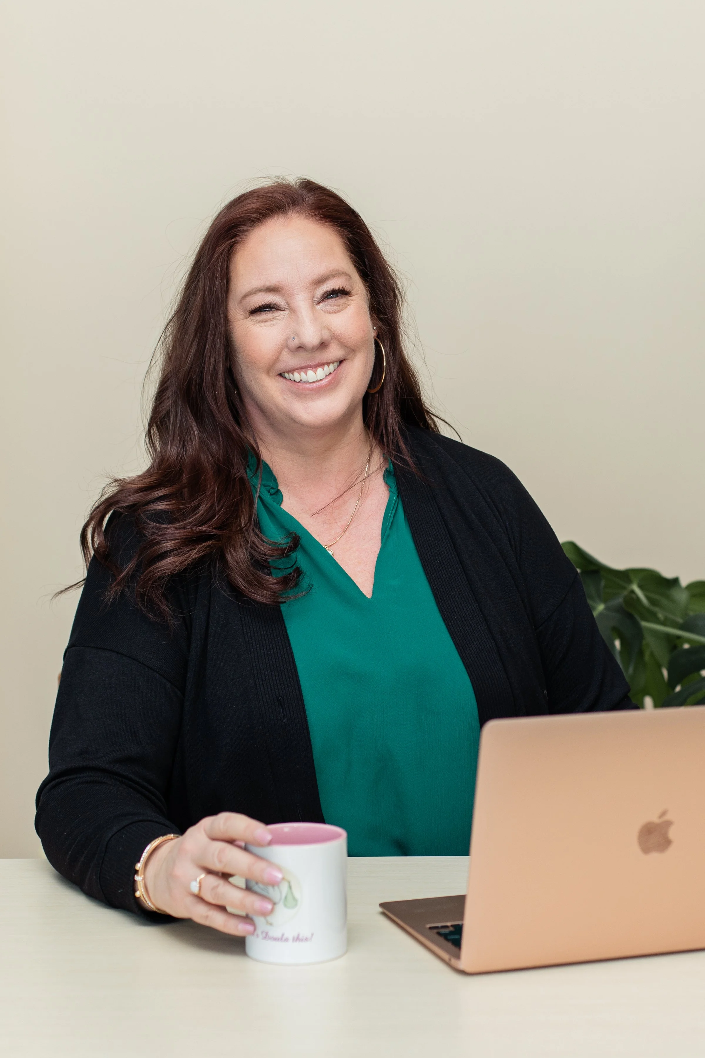 A woman with long wavy brown hair, wearing a green top and black cardigan, is sitting at a desk holding a white mug. She is smiling and looking at her laptop, which is on the desk in front of her. There is a plant partially visible behind her.