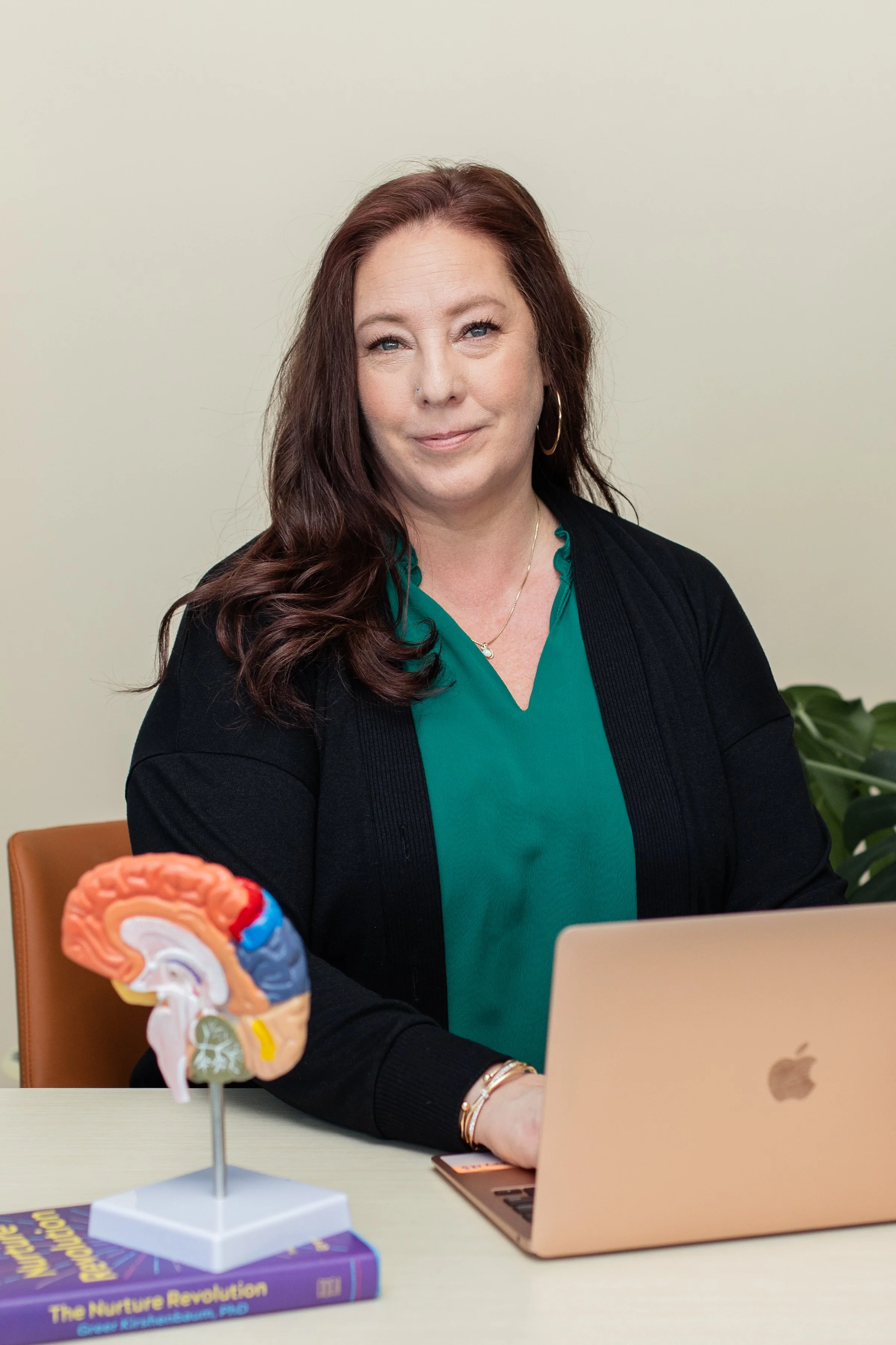 A woman with red hair, wearing a teal blouse and black cardigan, sitting at a table with a MacBook laptop and a human brain model. There is a book titled "The Nature Revolution" on the table and a potted plant in the background.