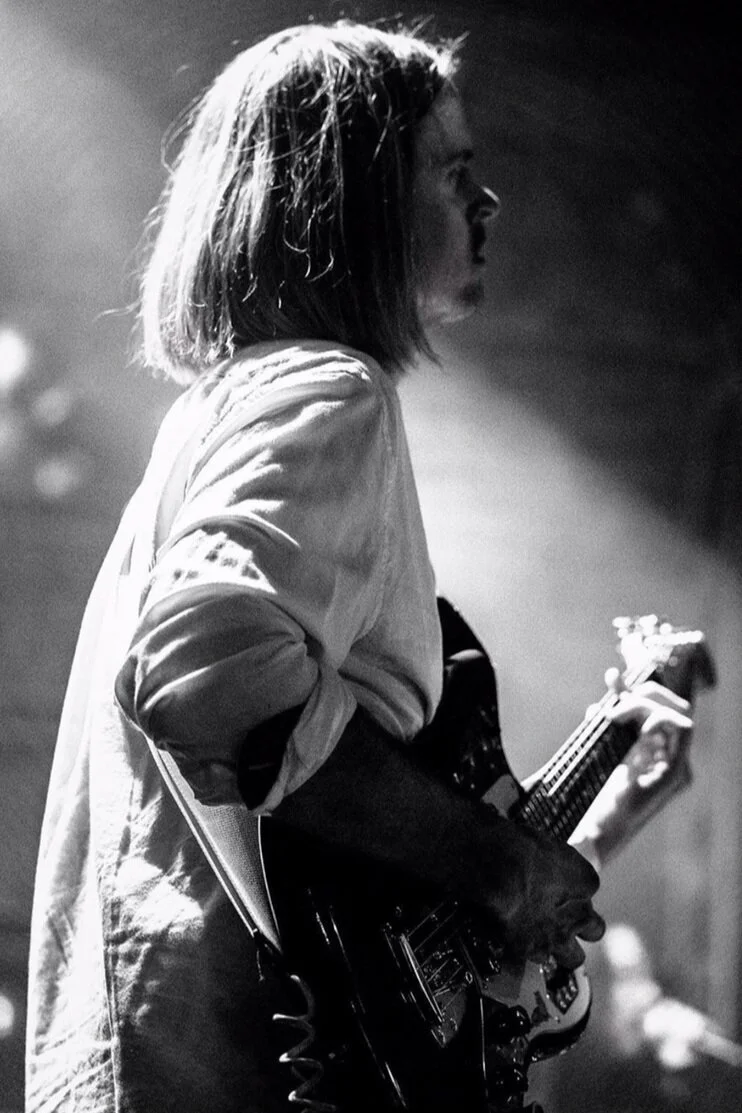 A woman with shoulder-length hair, wearing a light-colored jacket, playing an electric guitar on stage, in a black and white photo.