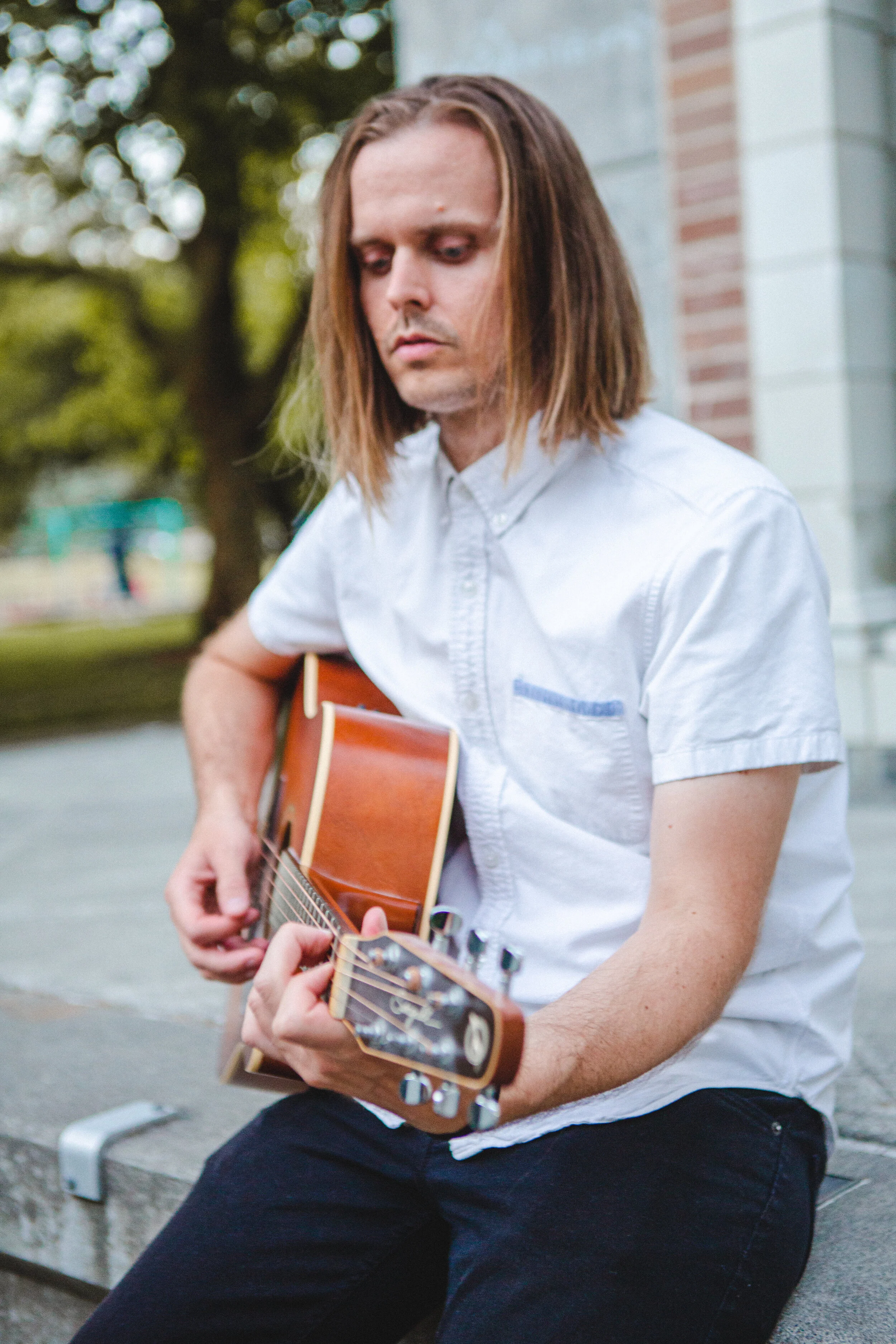 A man with shoulder-length brown hair playing an acoustic guitar outdoors, sitting on a concrete bench near trees and a brick building.