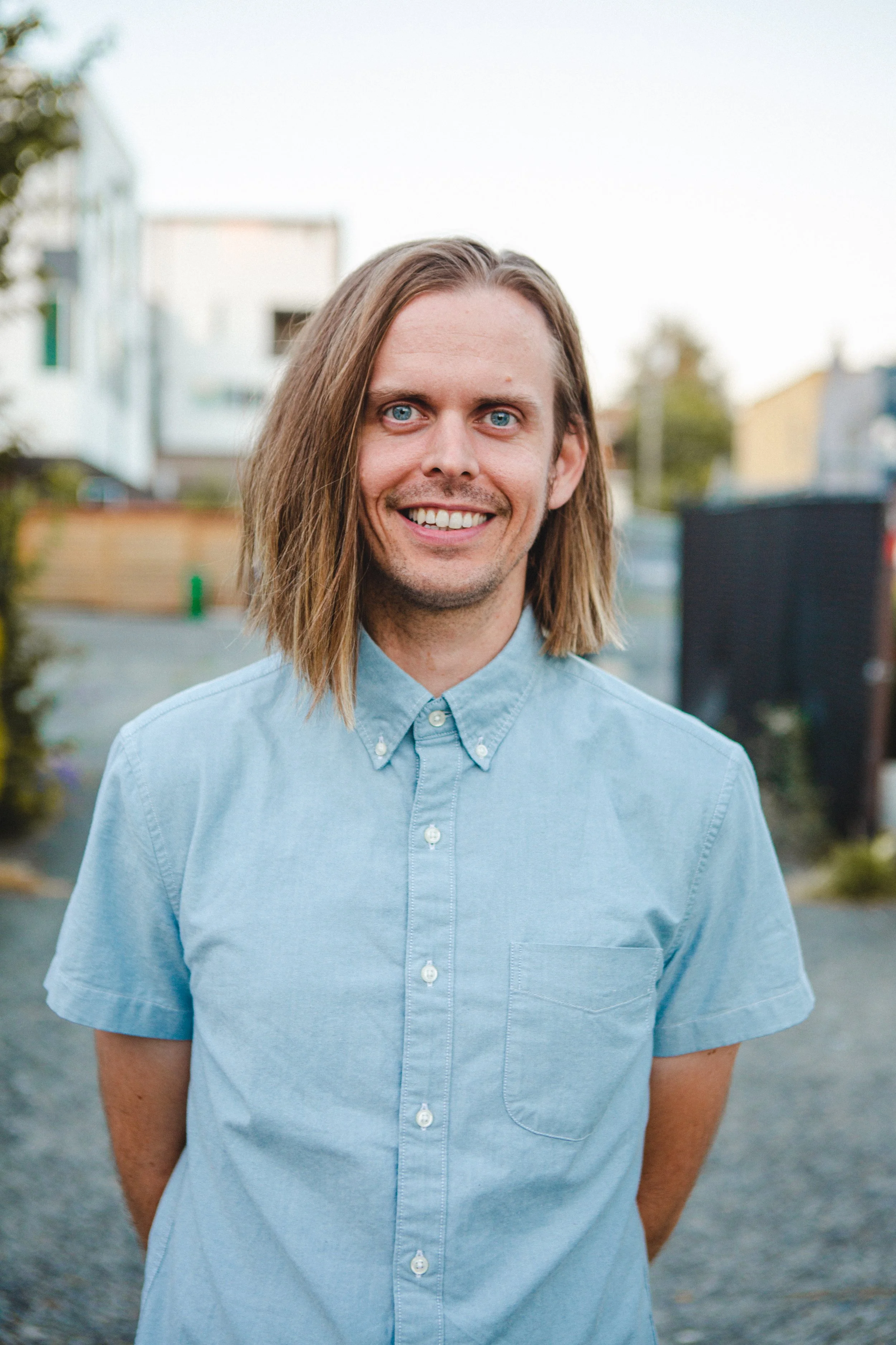 A young man with long blond hair, blue eyes, and a wide smile, wearing a light blue button-up shirt, standing outdoors.