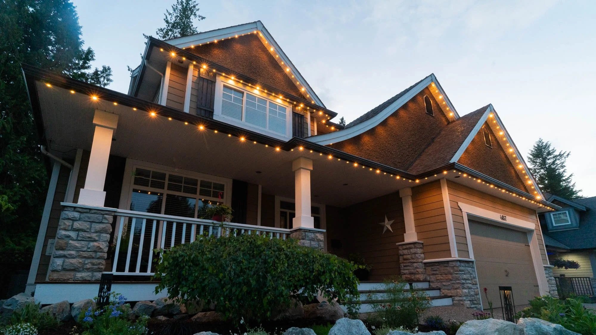 Large suburban home with a covered porch, stone accents, landscaped yard, and warm permanent Christmas lights glowing along the roofline at dusk