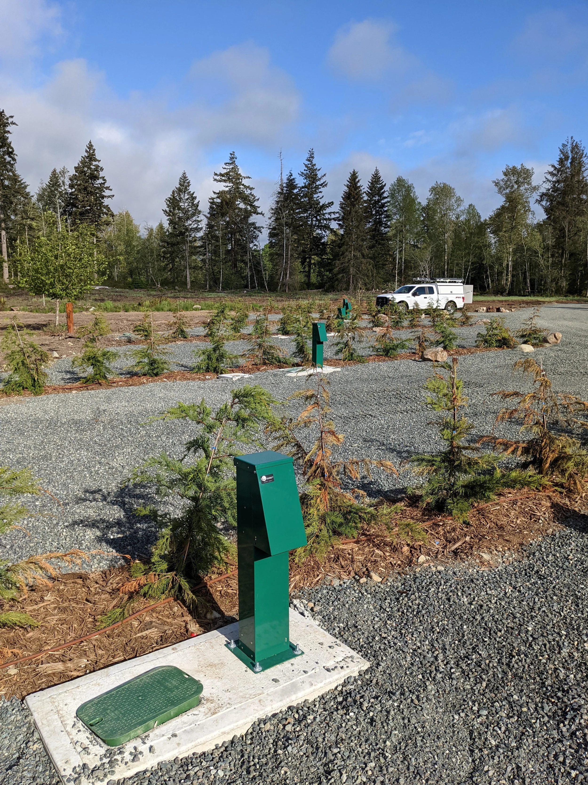 White utility pickup truck parked beside a forested camground with gravel landscaping, small trees, and green campground RV hookups