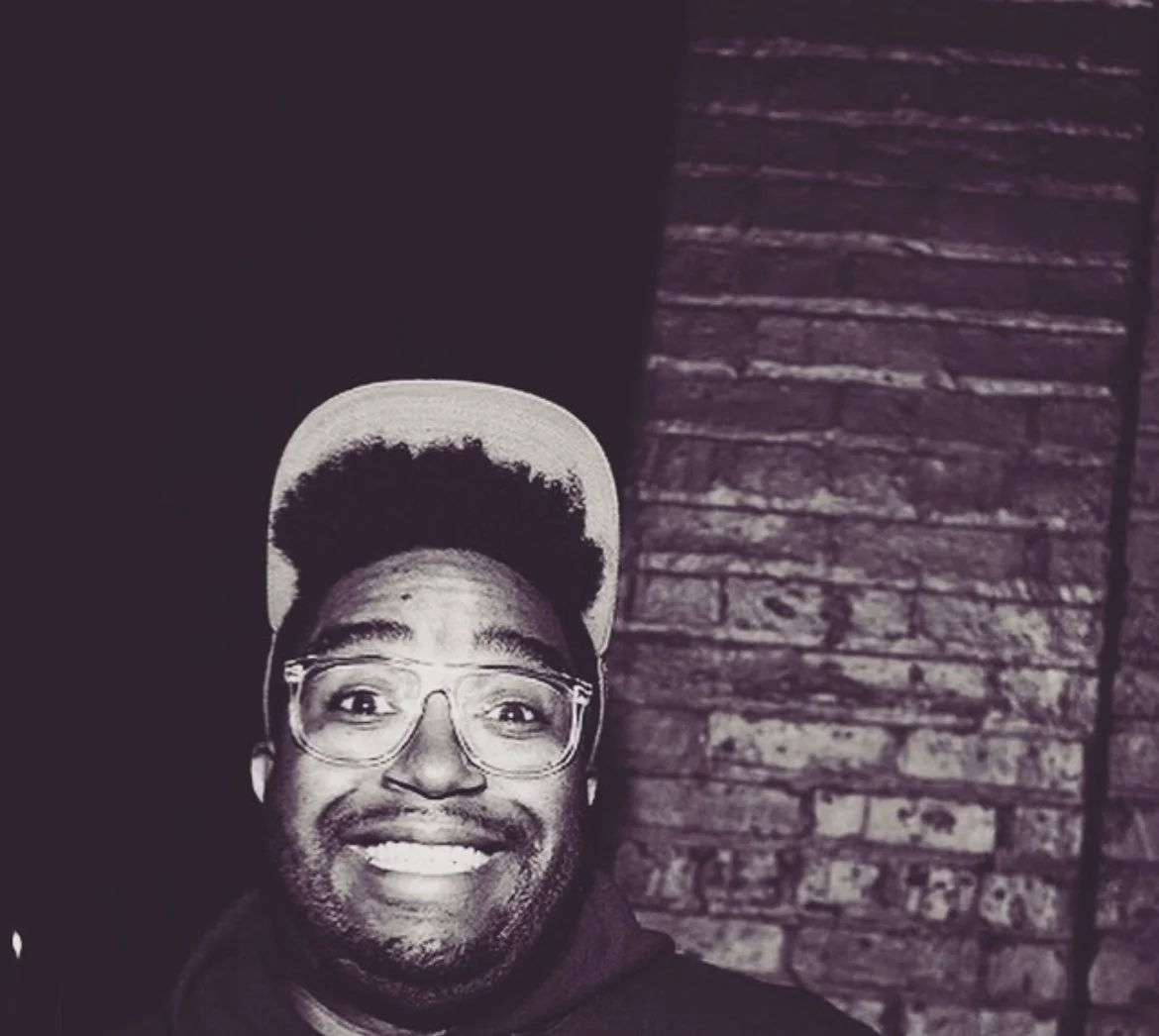 A smiling manwith curly black hair wearing glasses and a hat.