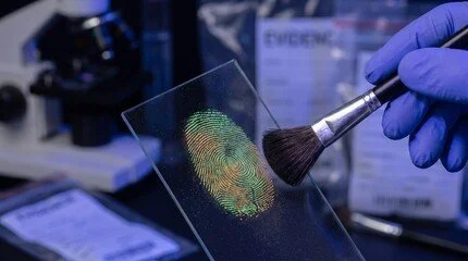 A person wearing blue gloves uses a brush to apply a fingerprint pattern onto a glass slide.