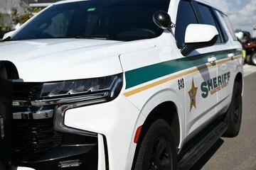 Front and side view of a white sheriff patrol vehicle with badge emblem and green and gold stripe.