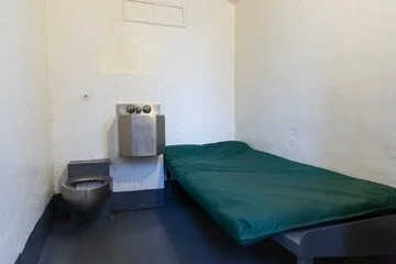 A small, sparse room with plain white walls, a simple bed with a dark green sheet, a water fountain mounted on the wall, and a black toilet in the corner.
