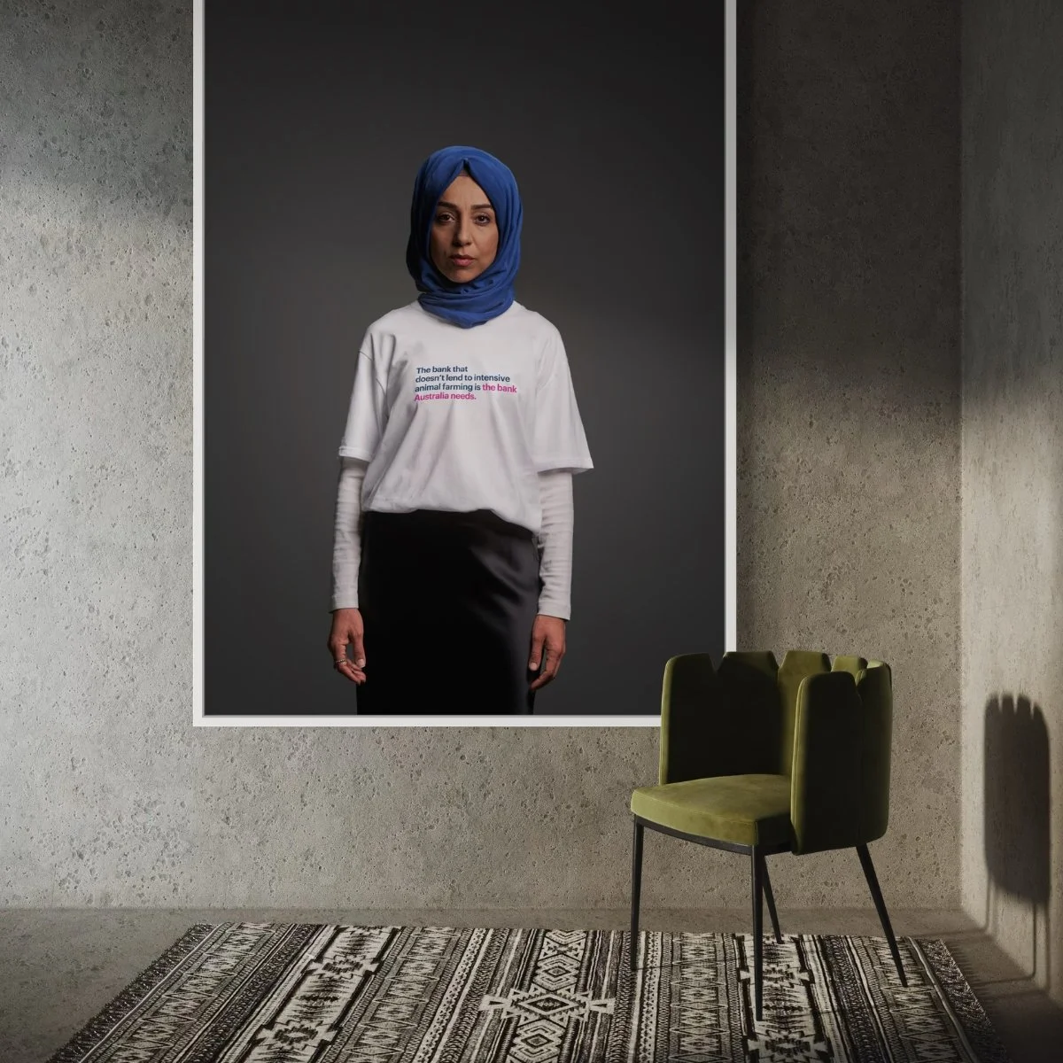 A gallery wall features a large portrait of a woman wearing a blue headscarf, white long-sleeve shirt, and dark skirt, with focus on the face. The shirt has text that reads, 'The bank that doesn't lend to intensive animal farming is the bank Australia needs.' An olive green chair and patterned rug are in front of the wall.