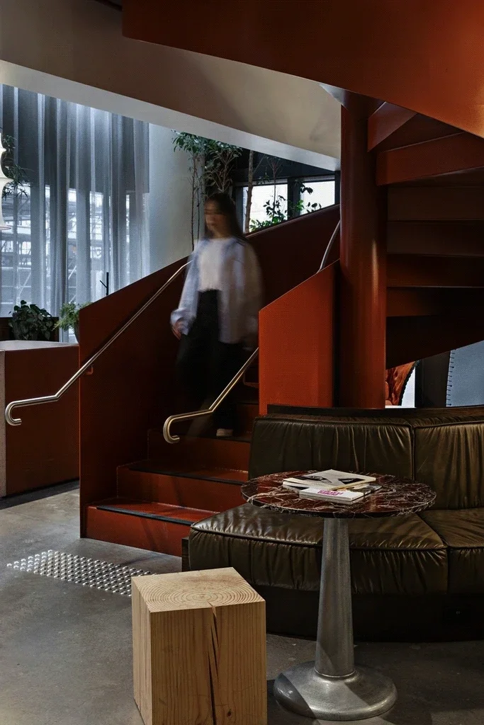 Interior of a modern building with a spiral staircase, a person walking downstairs, a seating area with a leather sofa, a small table with magazines, a wooden cube, and large windows with curtains and plants.