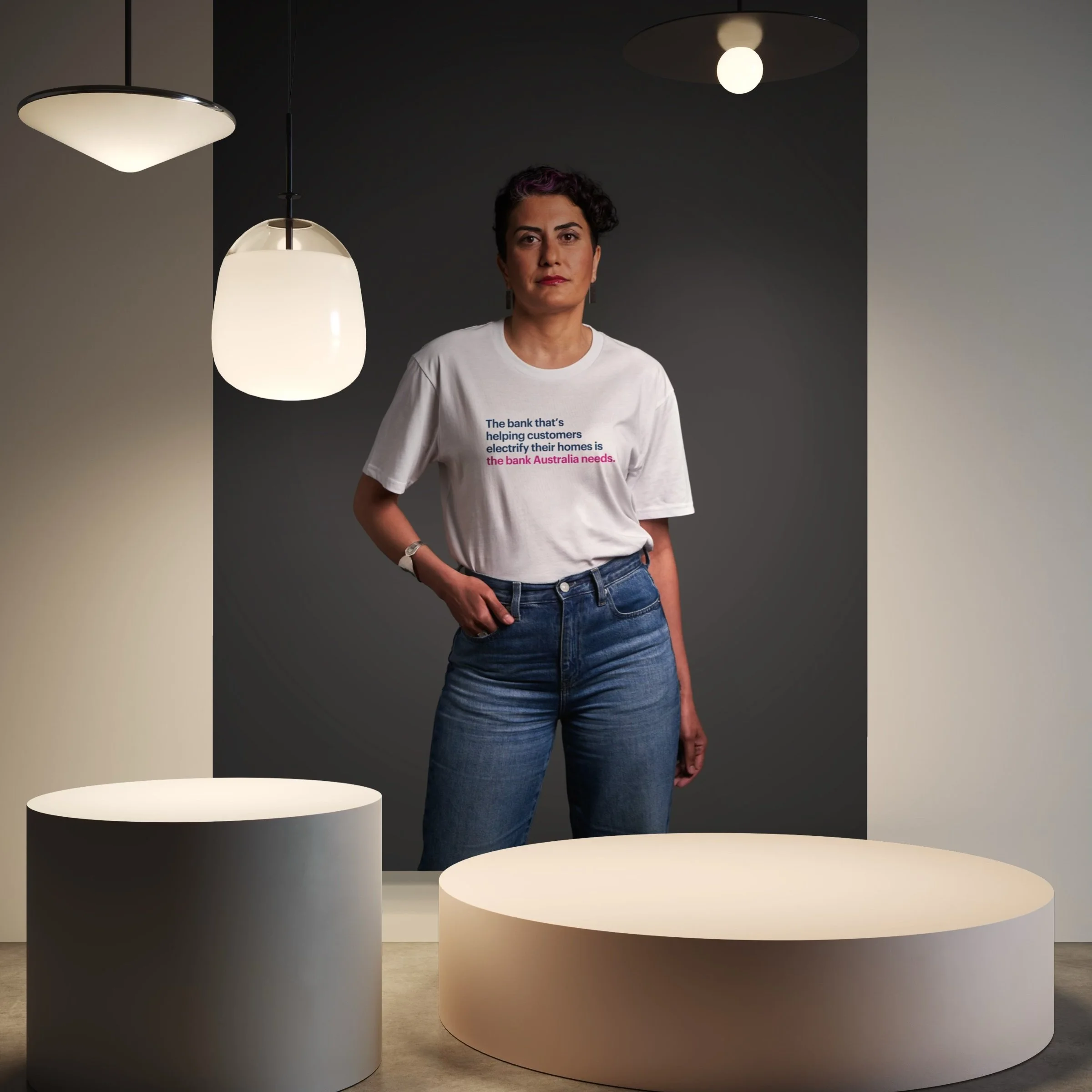 A woman with short curly hair standing in a modern room with round furniture and hanging lights. She is wearing a white T-shirt with multicolored text and blue jeans.
