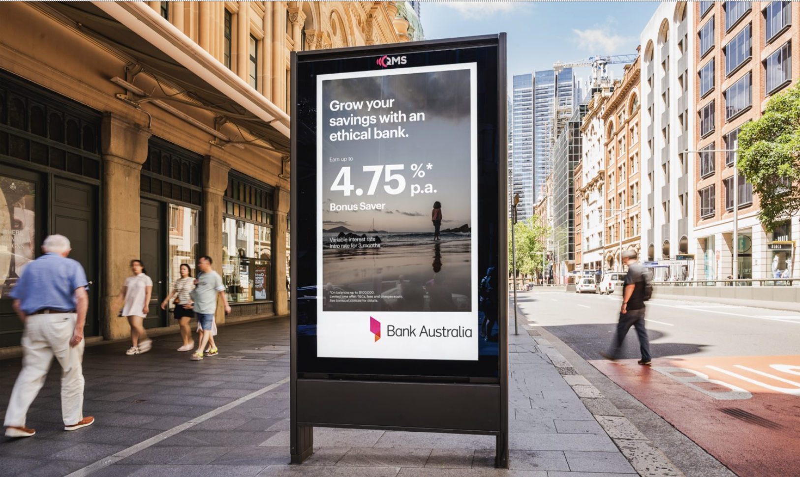 Digital billboard on city sidewalk advertising Bank Australia savings account with 4.75% interest rate, featuring a woman walking on the beach at sunset, with surrounding pedestrians and city buildings in the background.