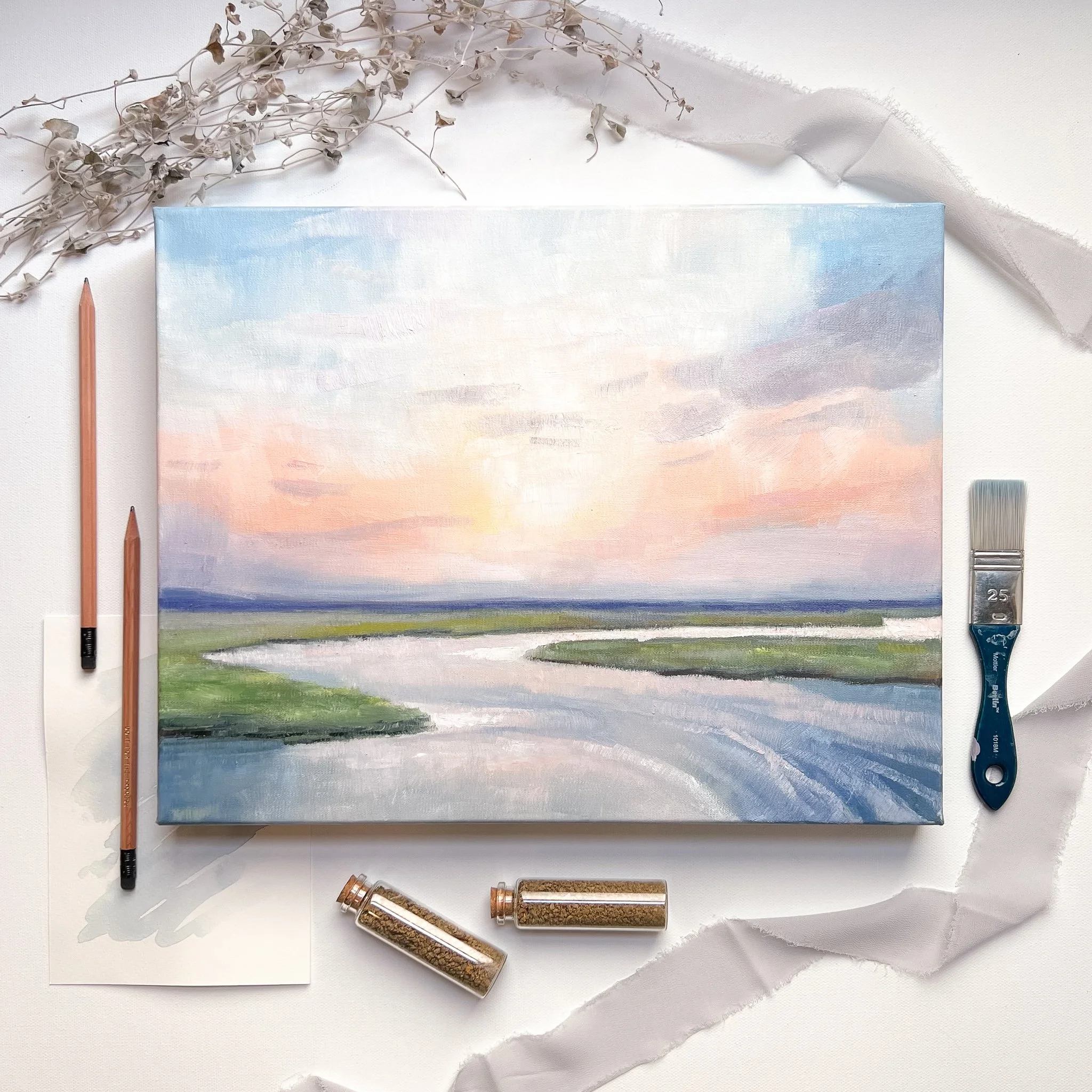 A landscape painting of a river at sunset with pink, purple, and blue sky, green grass, and flowing water, surrounded by art supplies including two pencils, a brush, and two small jars of paint or glitter.