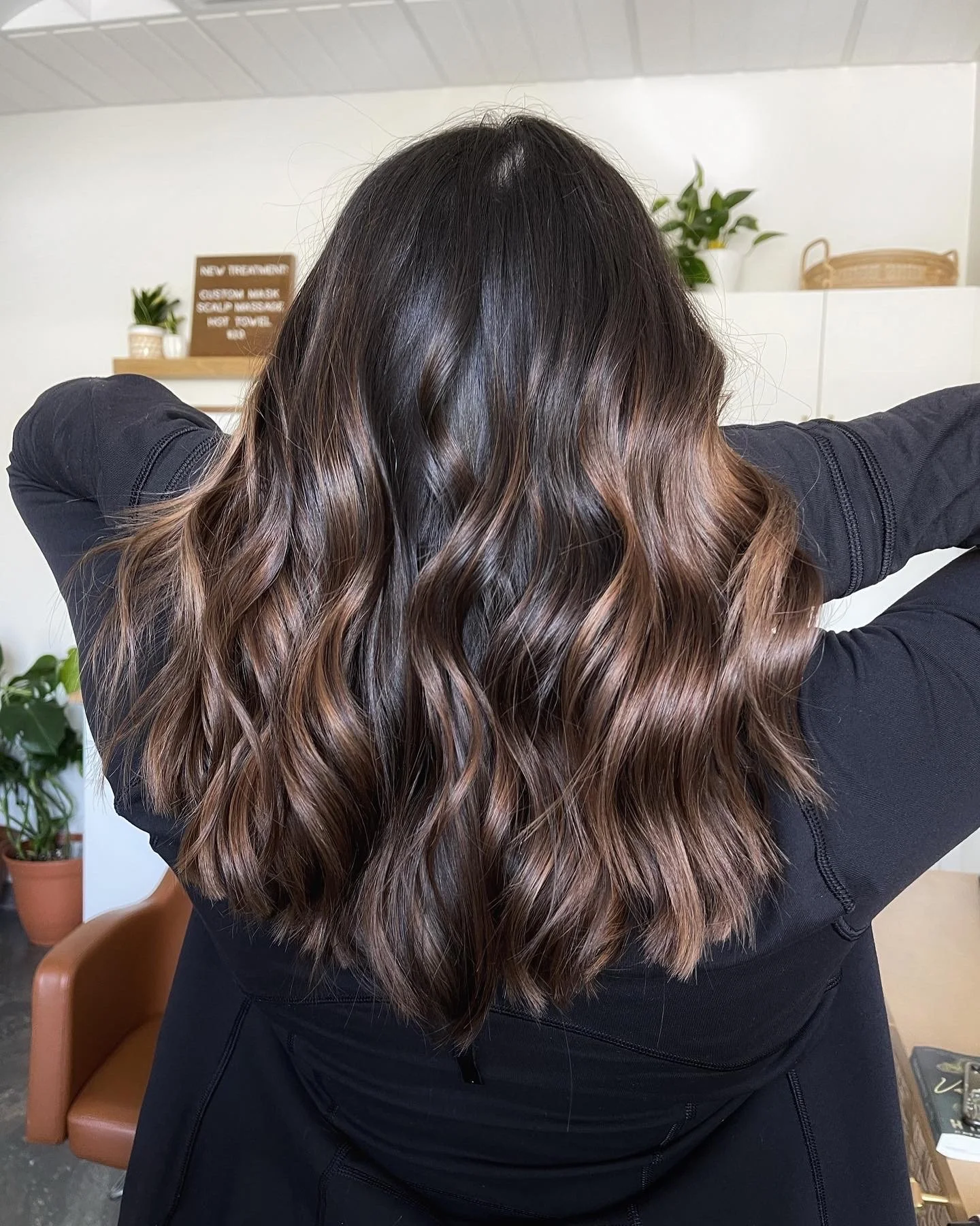 Back of a person with wavy, brown hair with subtle highlights, wearing a black top, in interior setting.