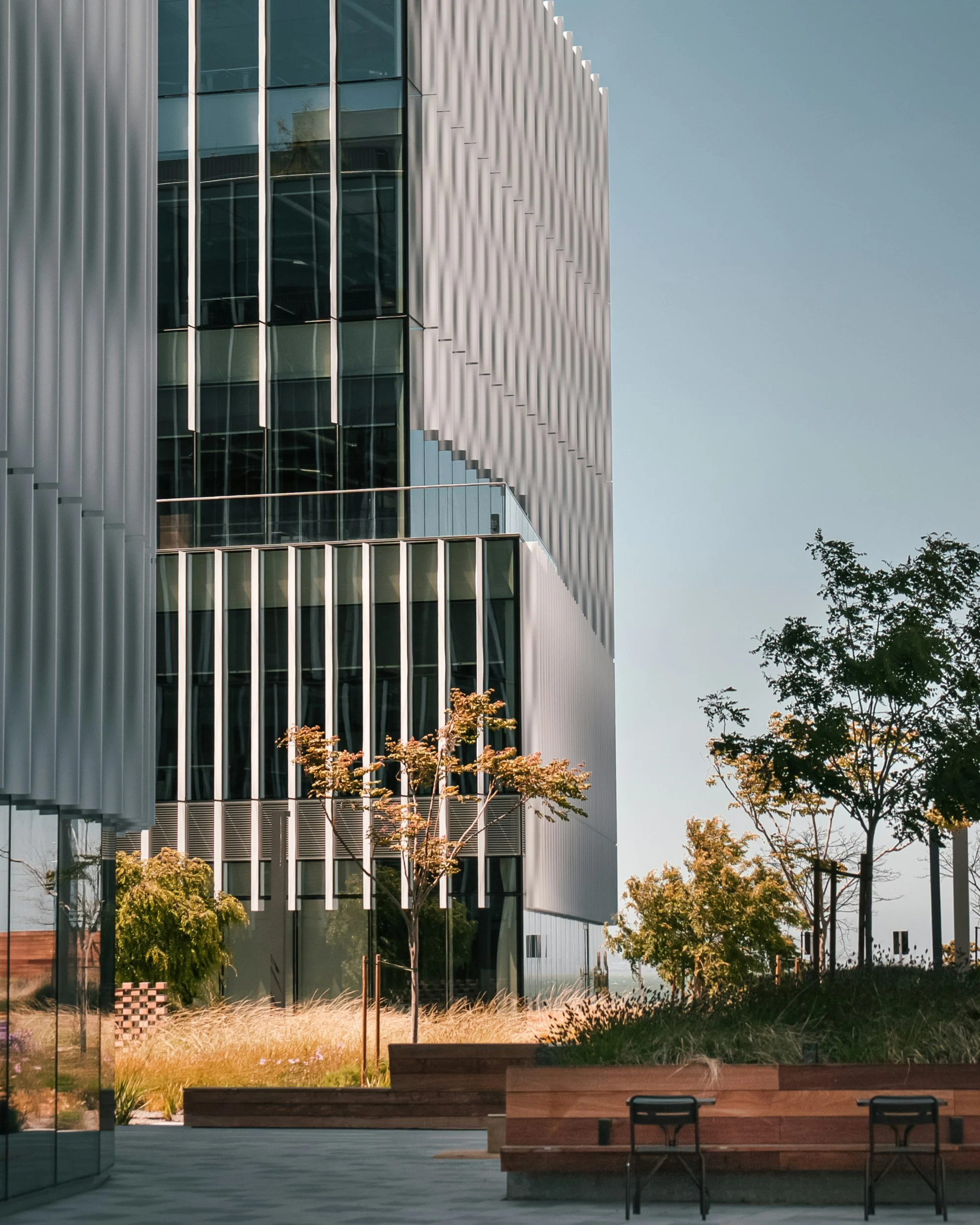 Modern building with vertical metallic and glass panels, trees in the foreground, and a clear sky.