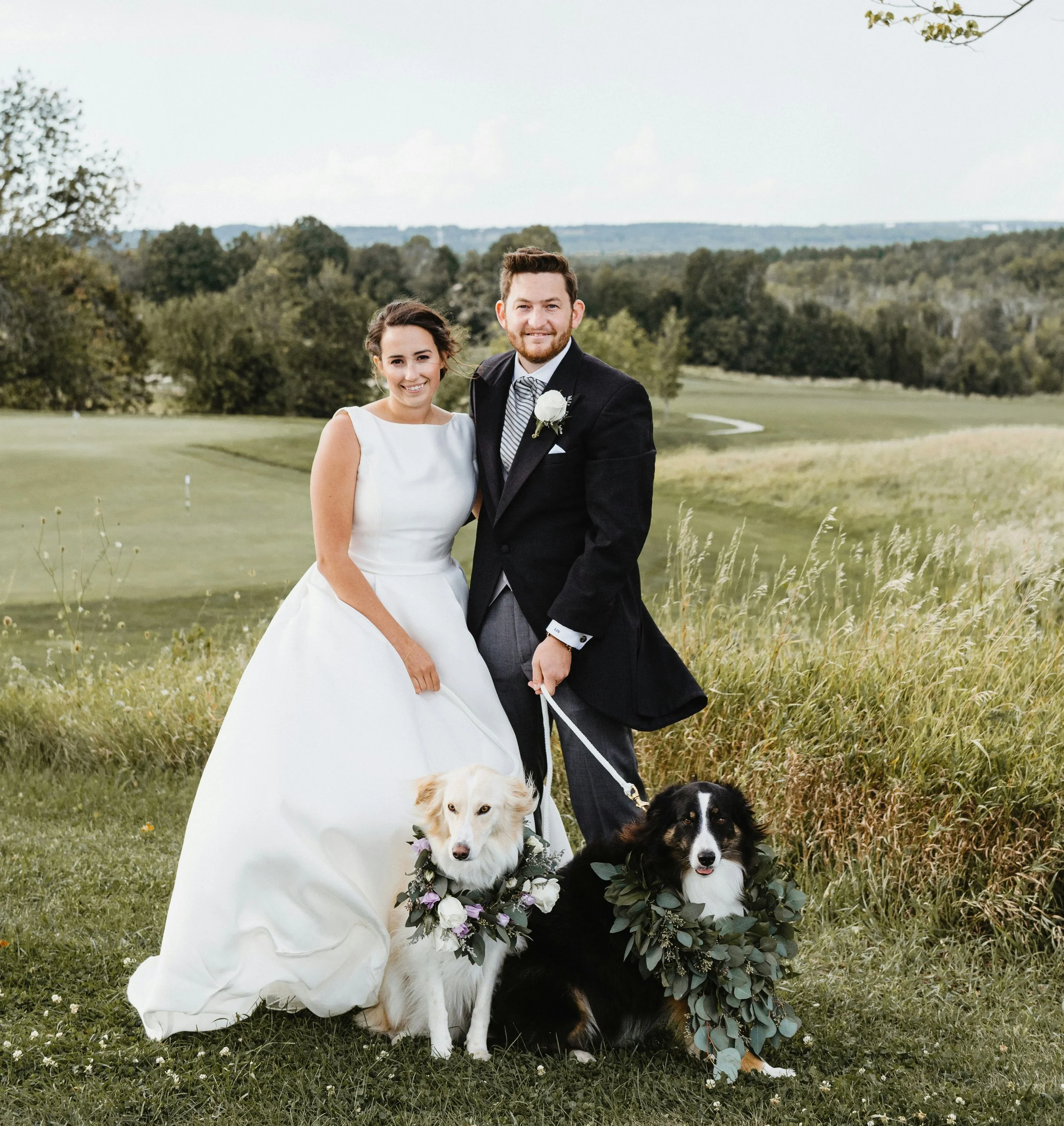 Bride and groom posing on a grassy hill with two dogs wearing elegant floral wreaths, utilizing professional dog wedding attendant services in Vancouver, BC.