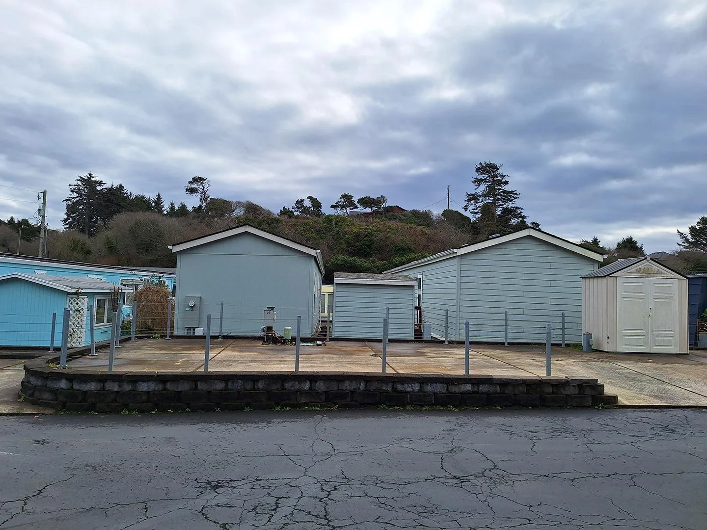 Photo of space available. Concrete slab, post & chain fence surrounding, storage shed with blue home behind.