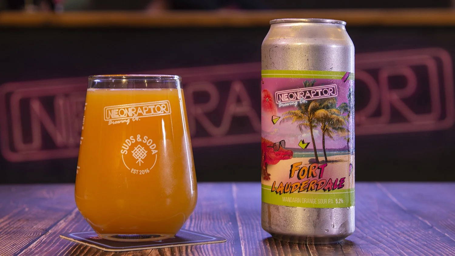 lifestyle product photography showing a glass and can of neon raptor craft beer on a wooden table with branded logo in the background