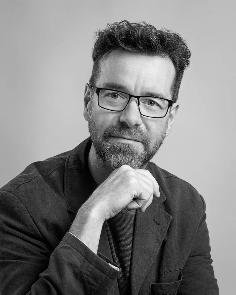 black and white portrait of roy mccready with glasses, a beard, and curly hair, resting his chin on his hand, looking at the camera
