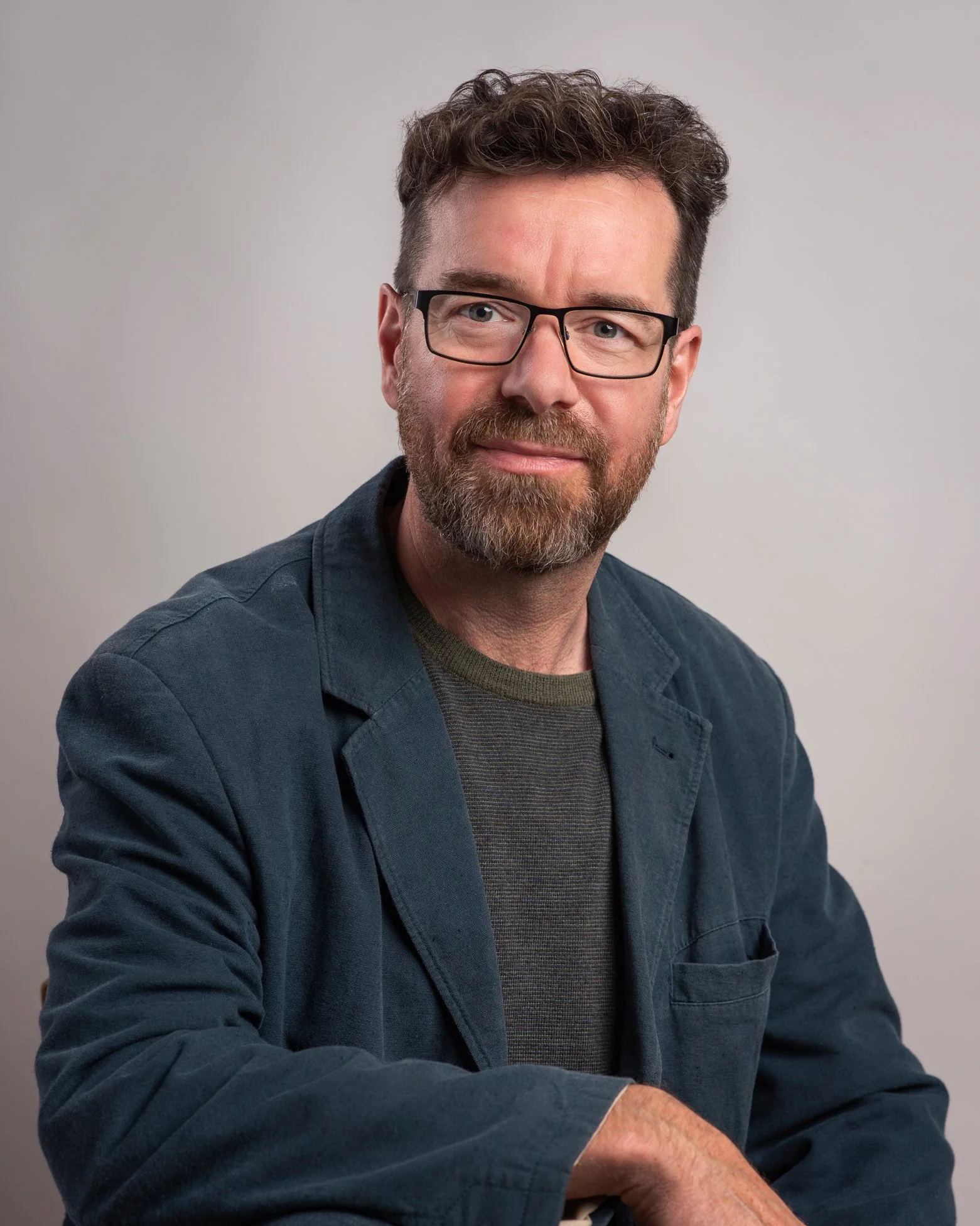 A branding portrait of roy mccready with glasses and a beard wearing a dark jacket and a gray shirt, sitting against a plain light-colored background.