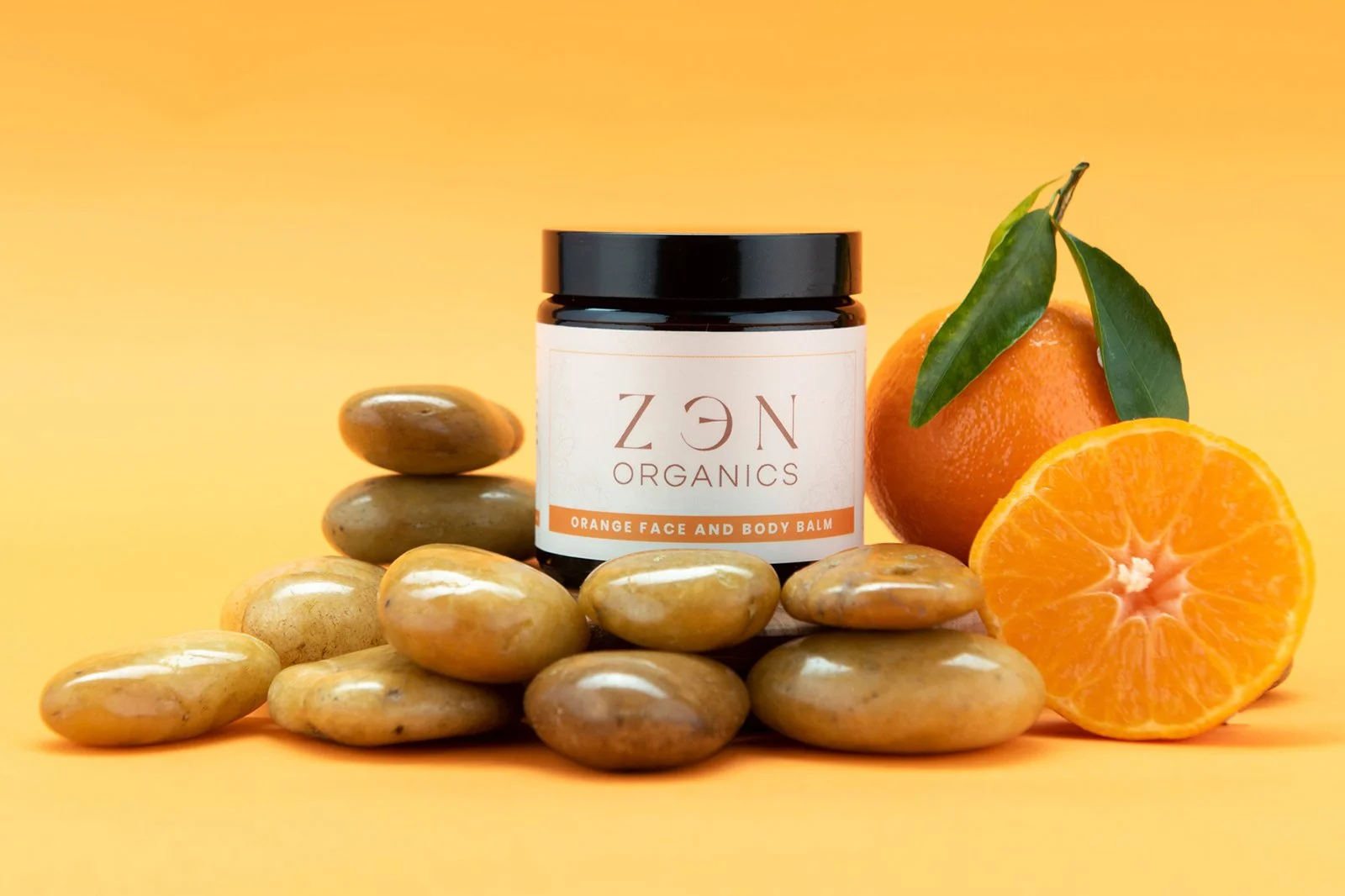 A jar of ZEN Organics face and body balm surrounded by smooth stones and a fresh orange with green leaves on a yellow background.