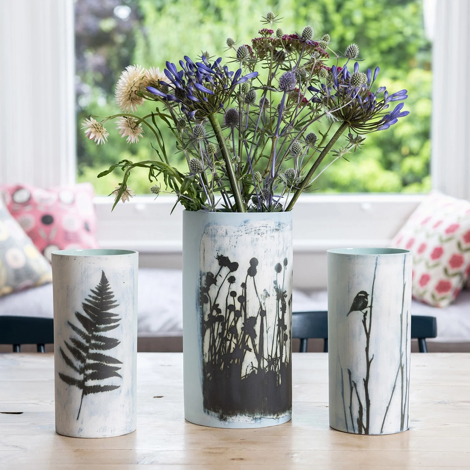 location lifestyle product photography showing 3 ceramic vases by fotoceramica on a wooden table with soft furnishing and window looking onto a garden the background