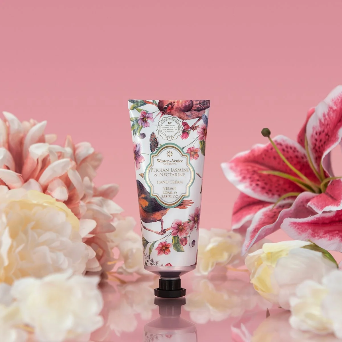 A tube of hand cream with floral and bird design on a pink background, surrounded by white and pink flowers.