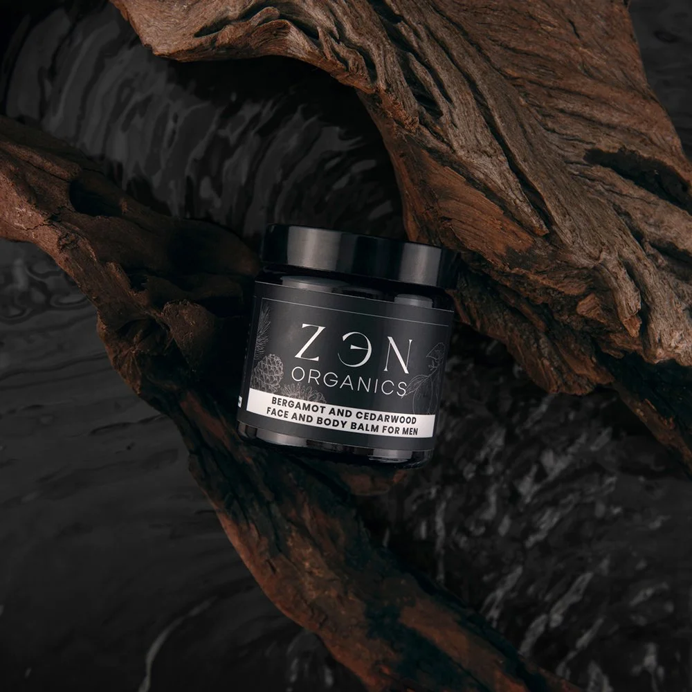 A black jar of Zen Organics Bergamot and Cedarwood face and body balm for men resting on a piece of dark, textured driftwood against a dark background.