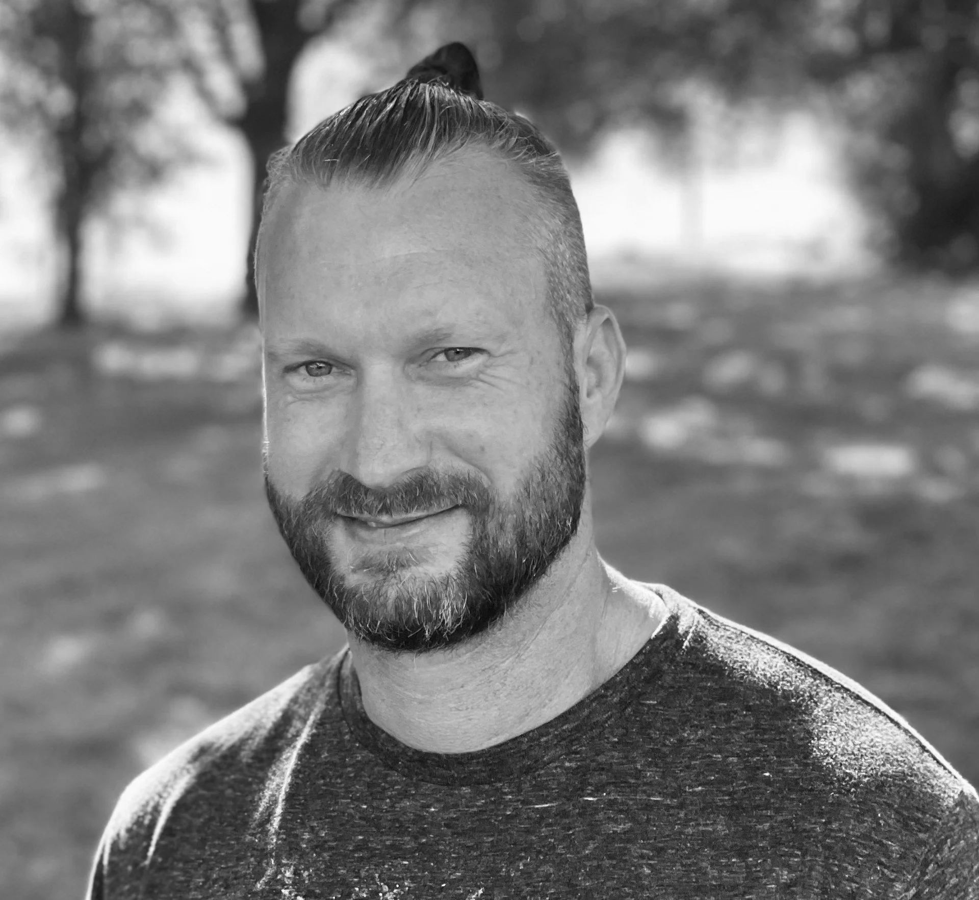 A black and white close-up photograph of a smiling man with tie-dye shirt in an outdoor park with trees in the background.