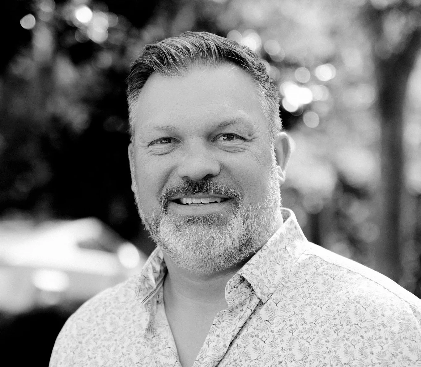 Black and white portrait of a smiling middle-aged man with a beard and short hair, wearing a patterned collared shirt, outdoors with blurred trees in the background.