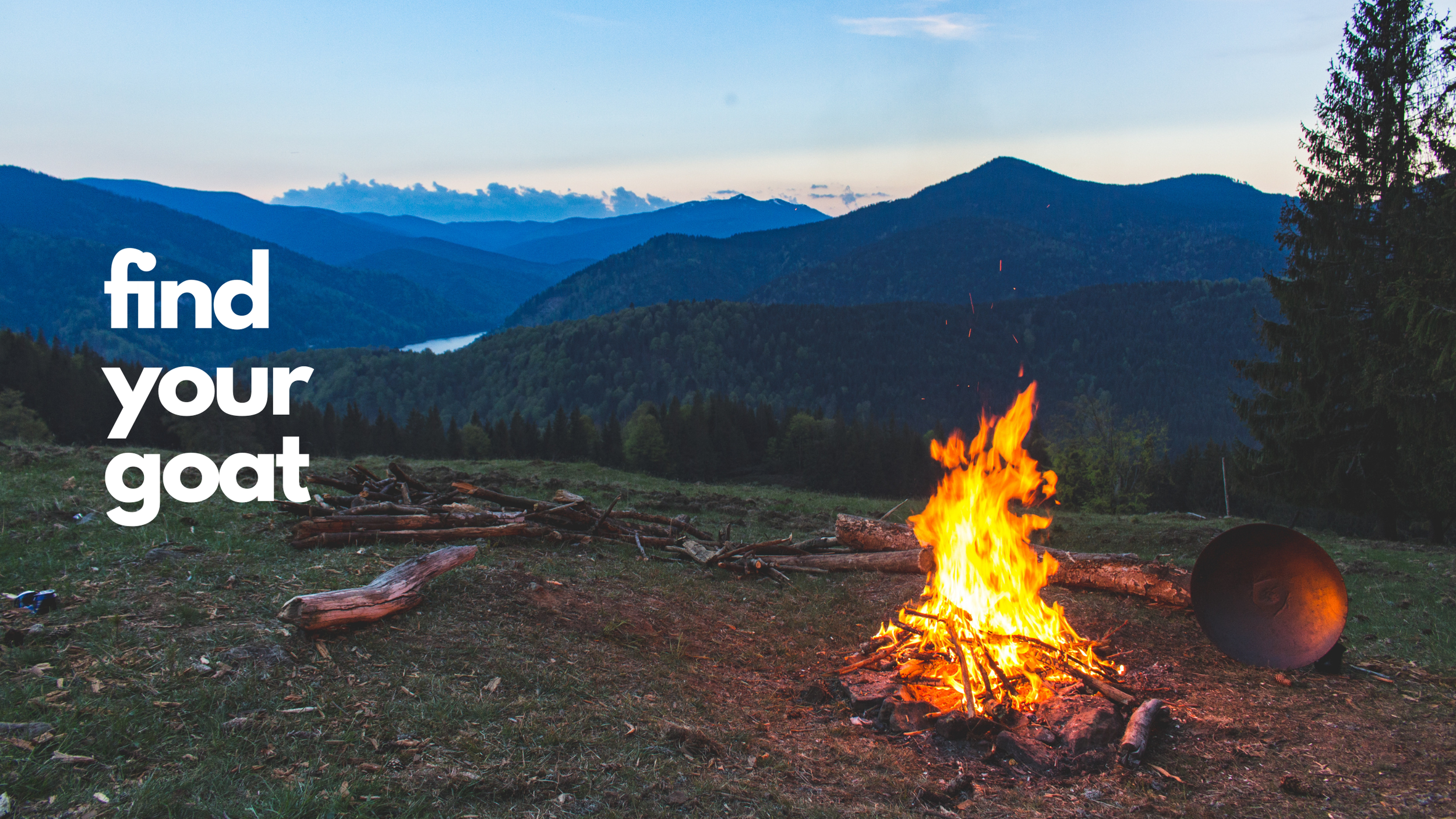 A campfire burning on a grassy hillside with a pile of logs, surrounded by trees, overlooking a mountain range and a river during sunset.