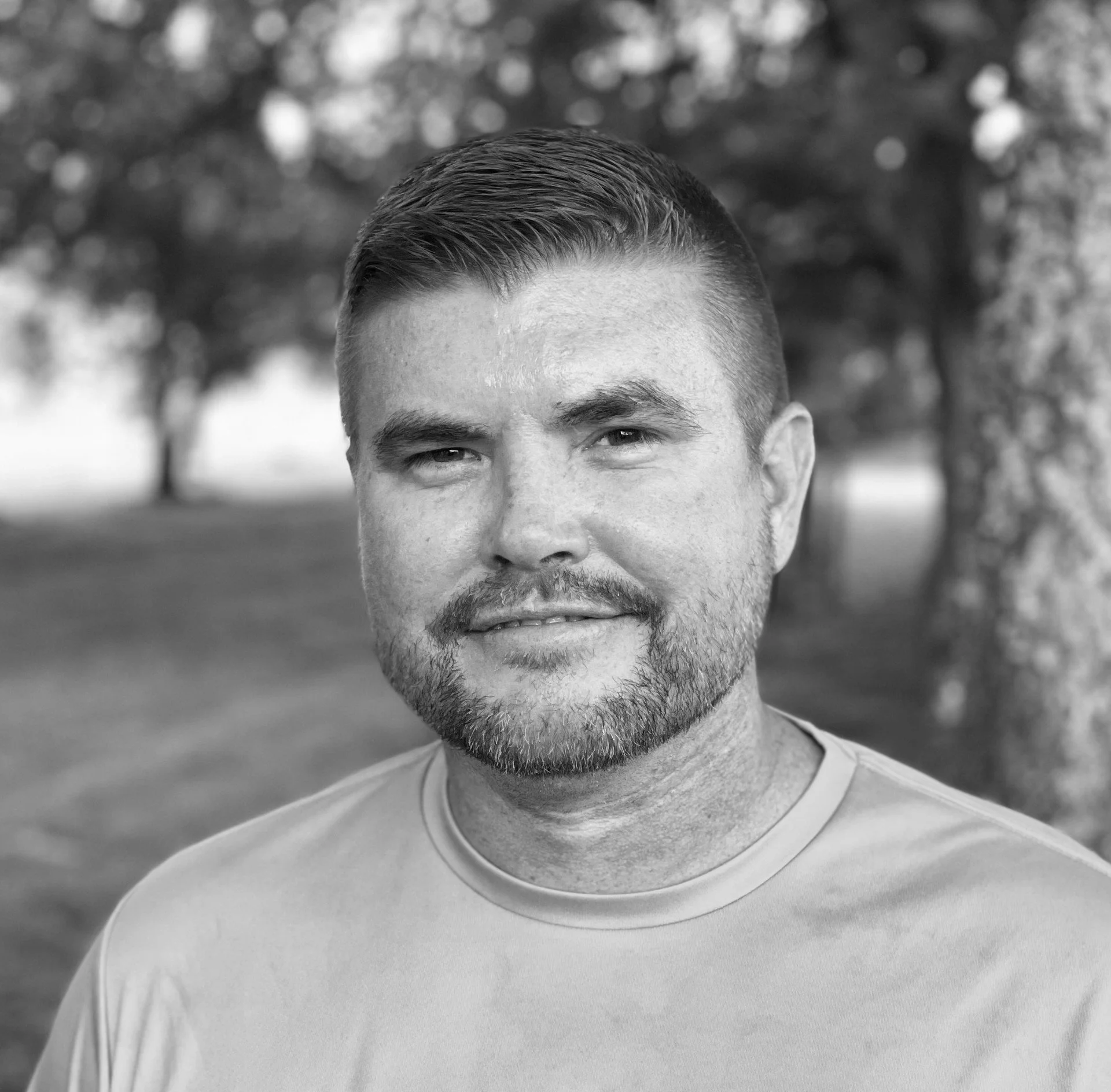 A black and white photo of a young man with short hair, slight beard, and mustache, wearing a T-shirt, standing outdoors with blurry trees in the background.