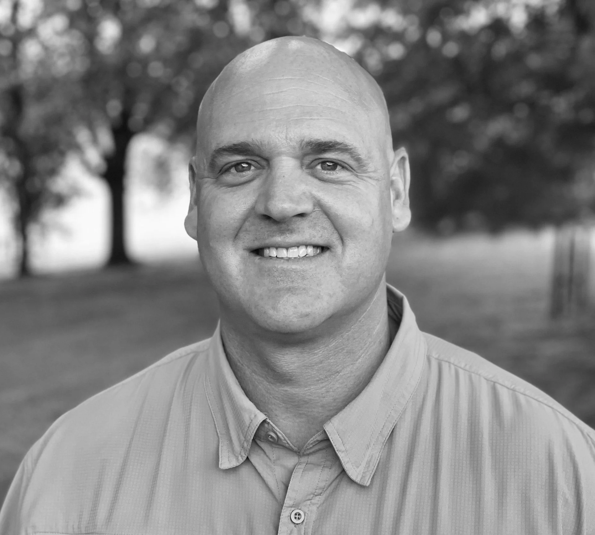 Black and white photo of a smiling man with a shaved head, wearing a collared shirt, standing outdoors with blurred trees in the background.