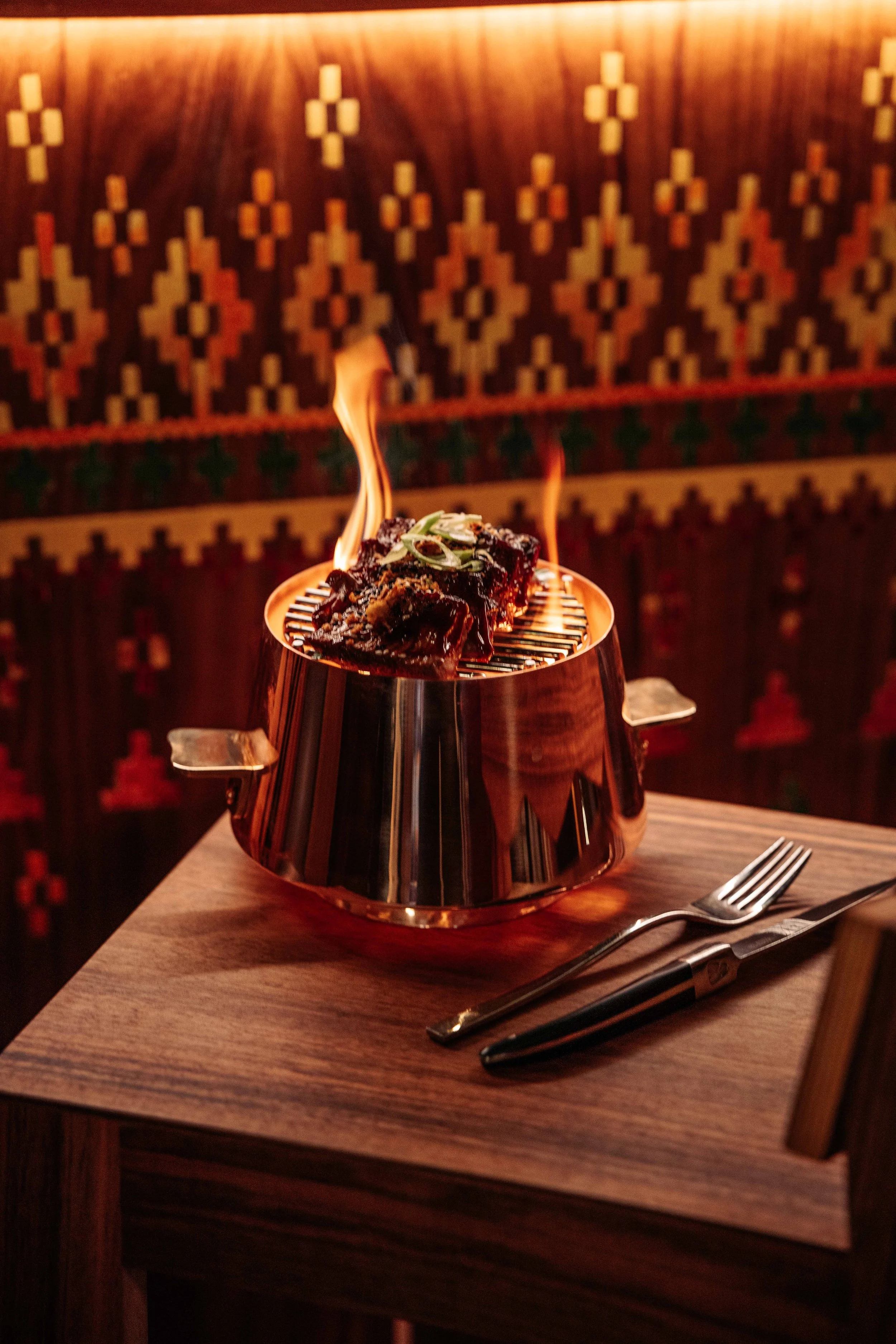 A rustic table with flaming meat dishes, set against a wooden background.