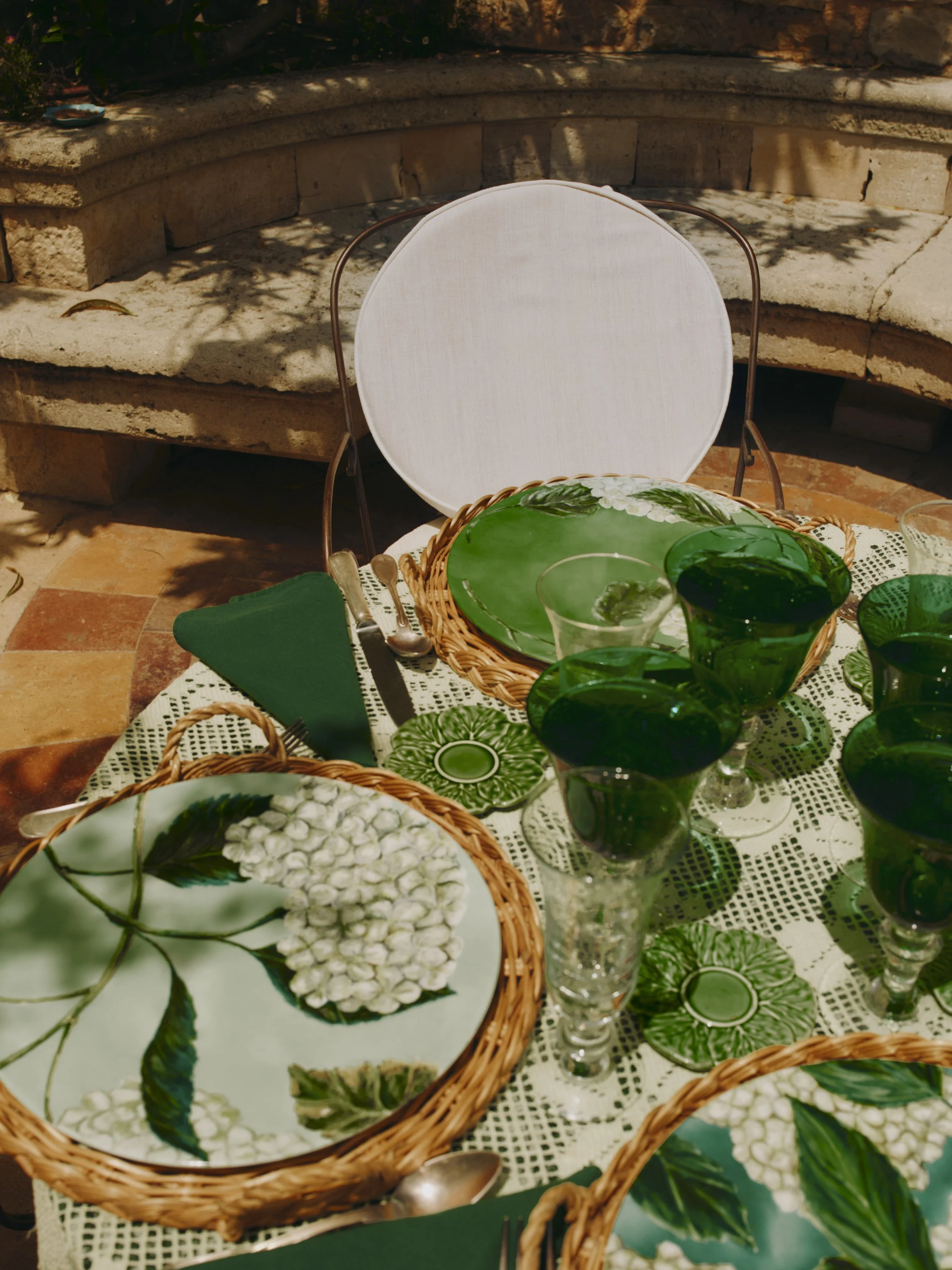 Table dressée pour un repas en extérieur avec des assiettes à motif floral, des verres verts, des couverts avec une serviette bleue et des petits sous-verres verts, ensoleillée avec ombres de feuilles.