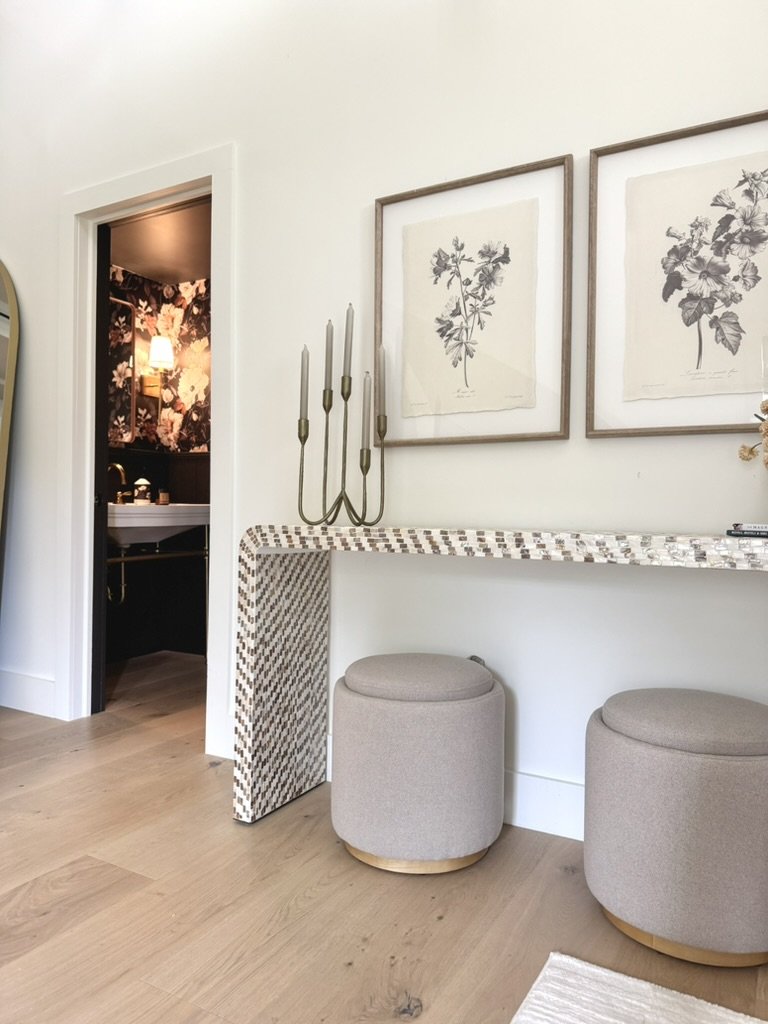 Interior of a room with two beige ottomans, a decorative table with a mirror design, a wall-mounted candle holder, framed botanical artwork, and a doorway leading to a bathroom with dark floral wallpaper, a sink, and a lamp.