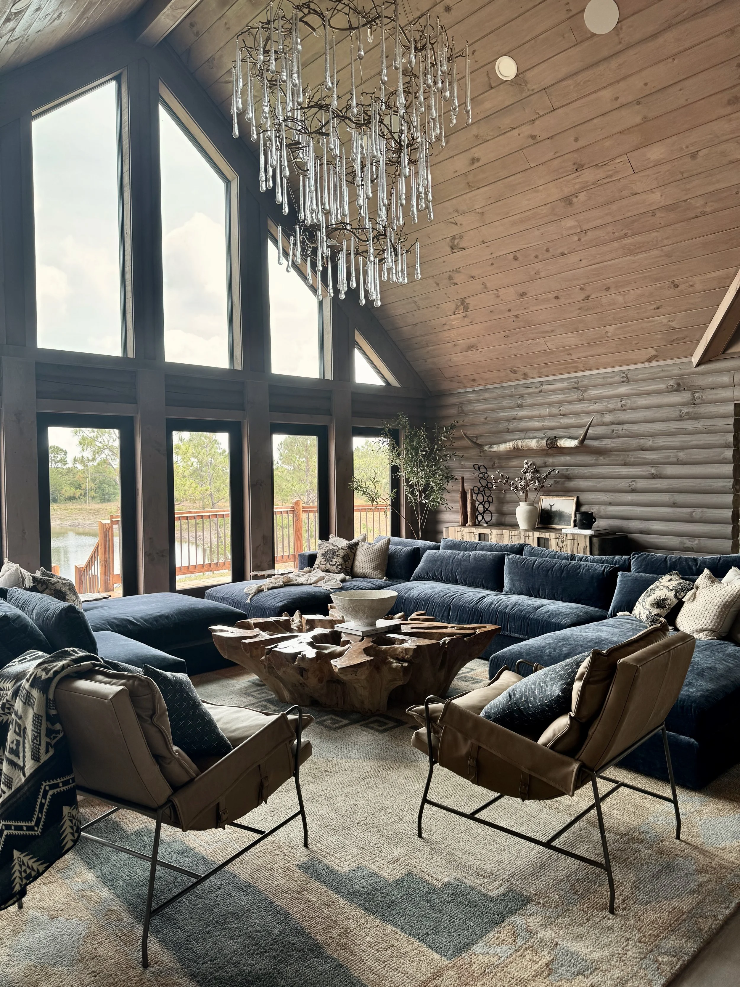 Inside a cozy living room with a vaulted ceiling, large windows revealing a lakeside view, a crystal chandelier hanging from the ceiling, a wooden coffee table, a large blue sectional sofa with assorted pillows, two tan chairs with metal frames, and 