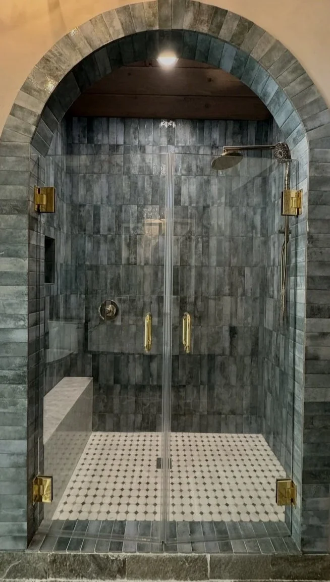 A glass shower enclosure with dark gray tiled walls, a built-in seat, and brass hardware. The shower has a rain showerhead and a recessed shelf, set within a stone archway.