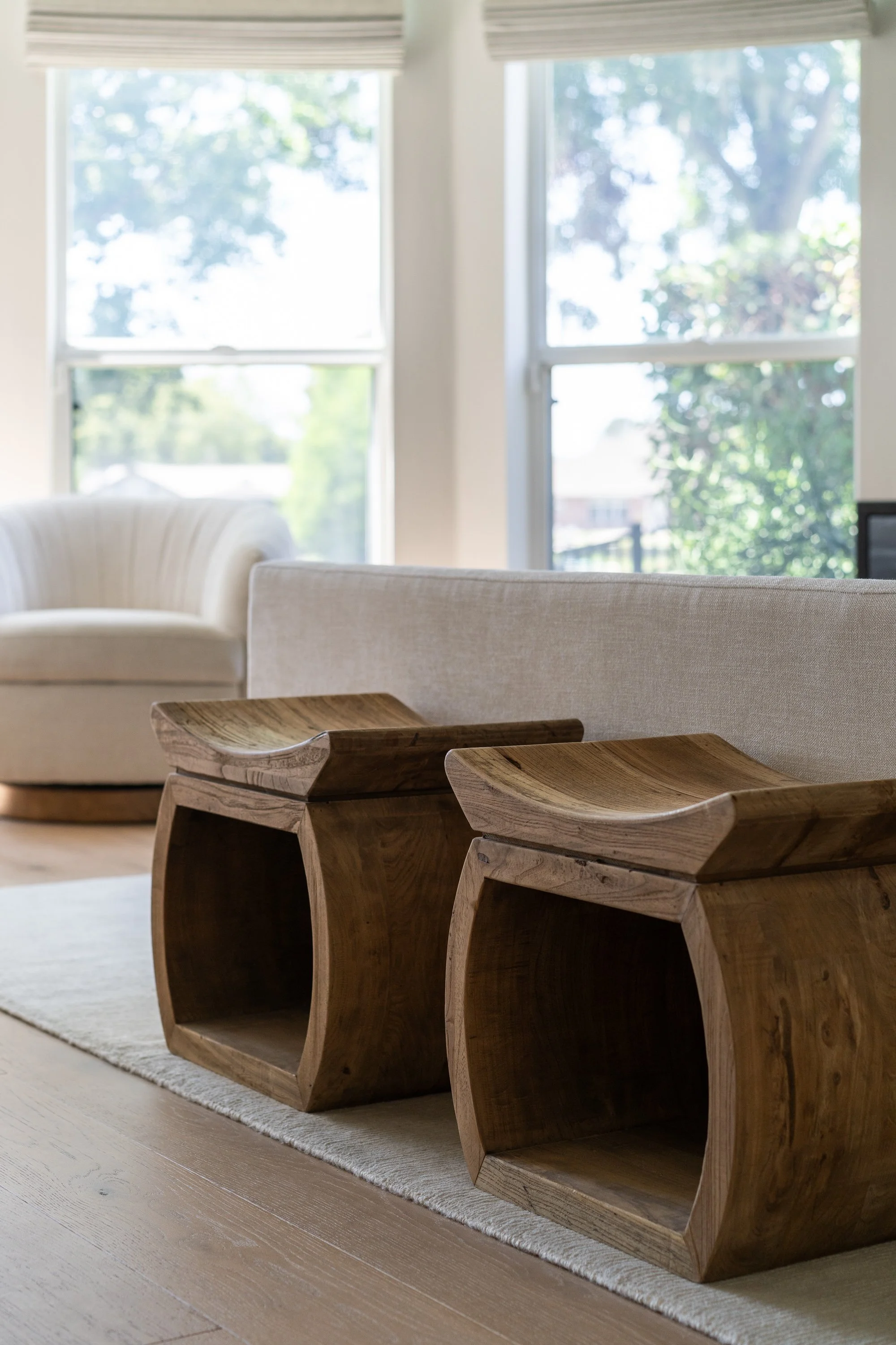 Living room with two wooden side tables, beige sofa, and large windows showing greenery outside.