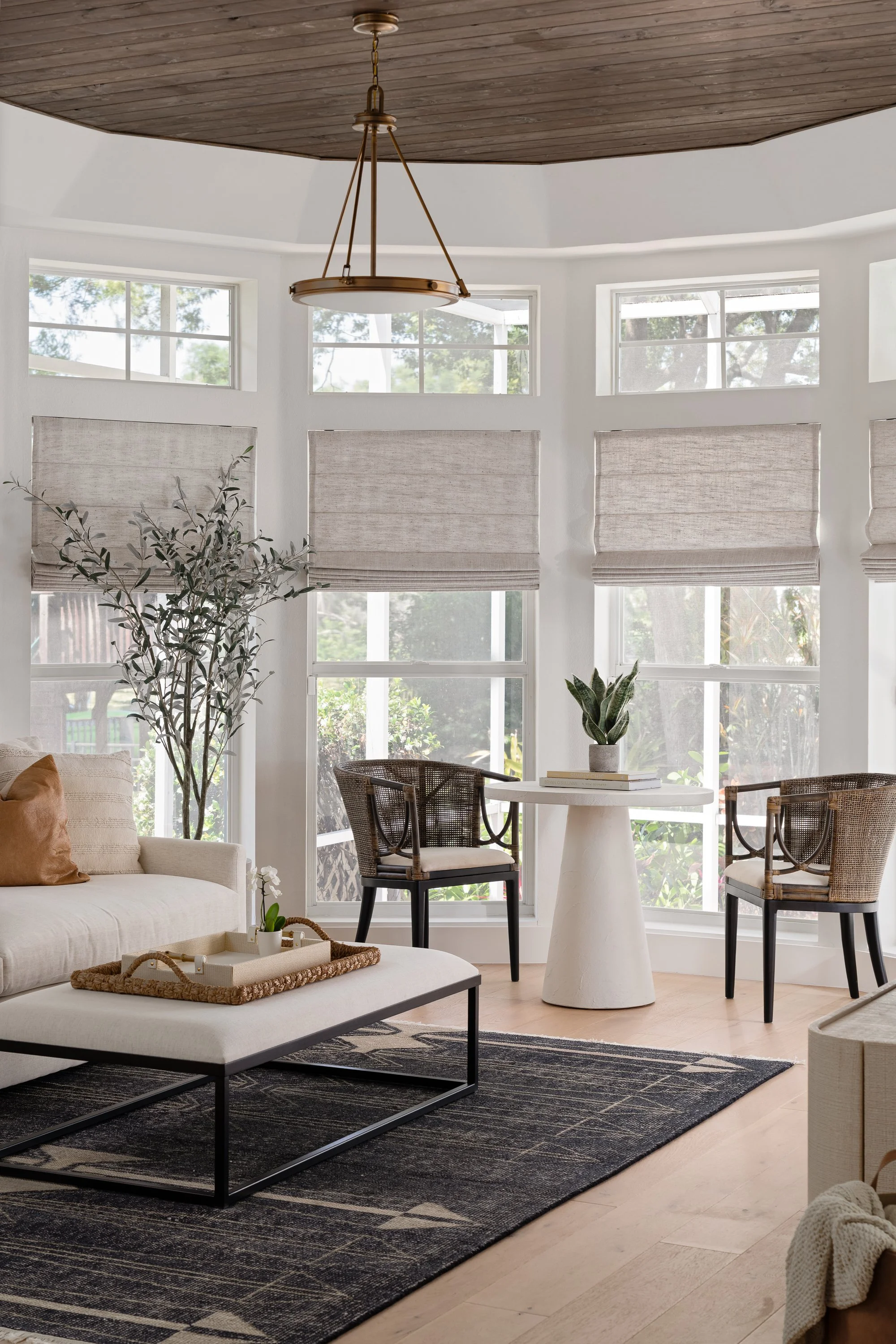 Bright living room with large windows, a beige sofa, a black and white rug, a white round table, two wicker chairs, a potted plant, and window shades.