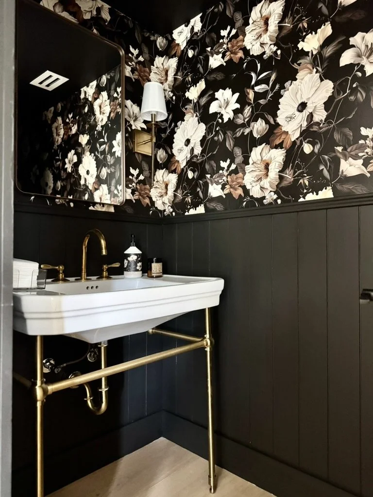 A bathroom with a floral patterned wallpaper, a white sink with gold fixtures, a black soap dispenser, a small bottle, a mirror, a wall sconce, and dark paneling on the lower part of the wall.