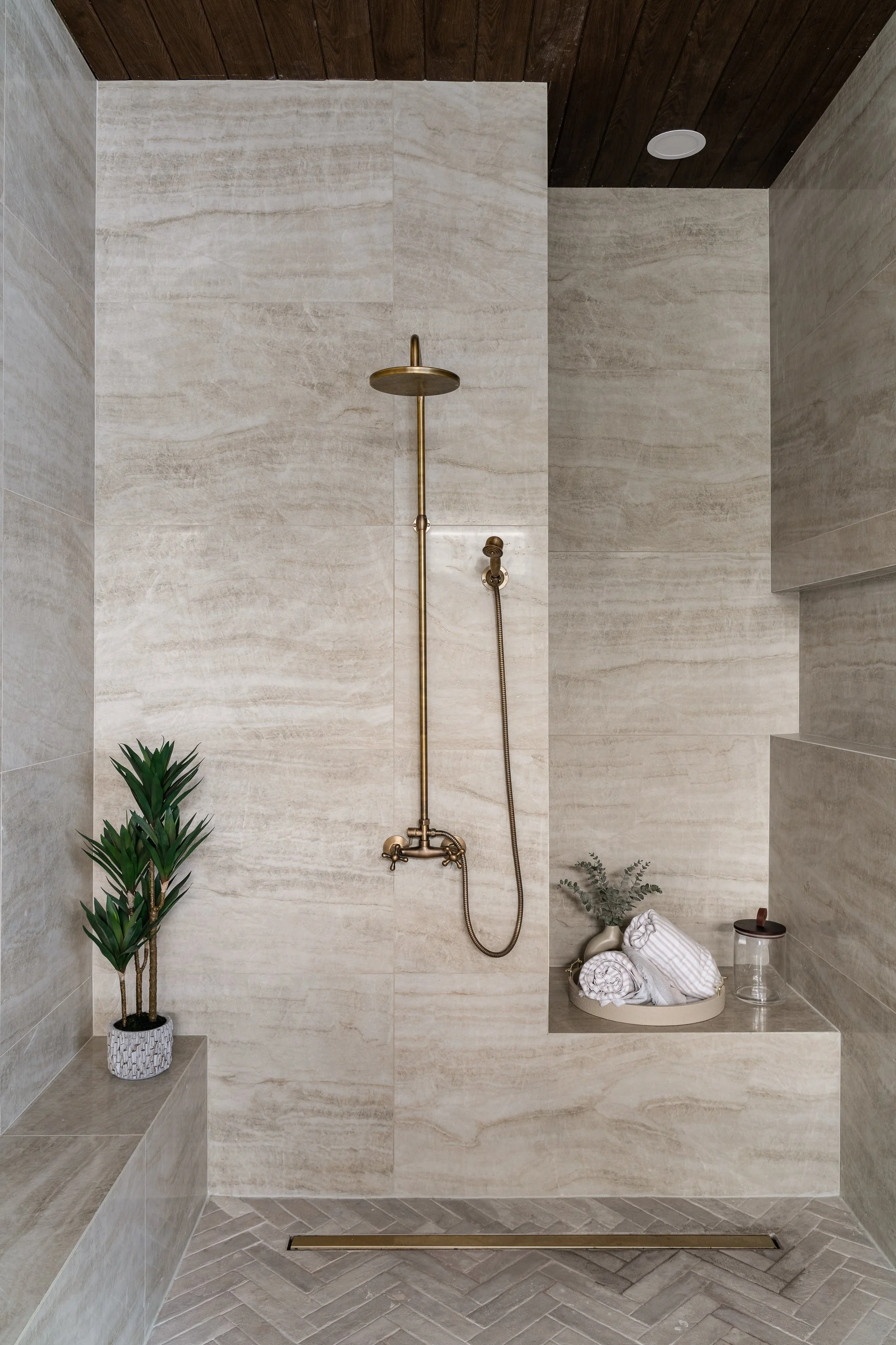A modern walk-in shower with beige marble tiles, a brass showerhead, and a built-in seating ledge decorated with towels and plants.
