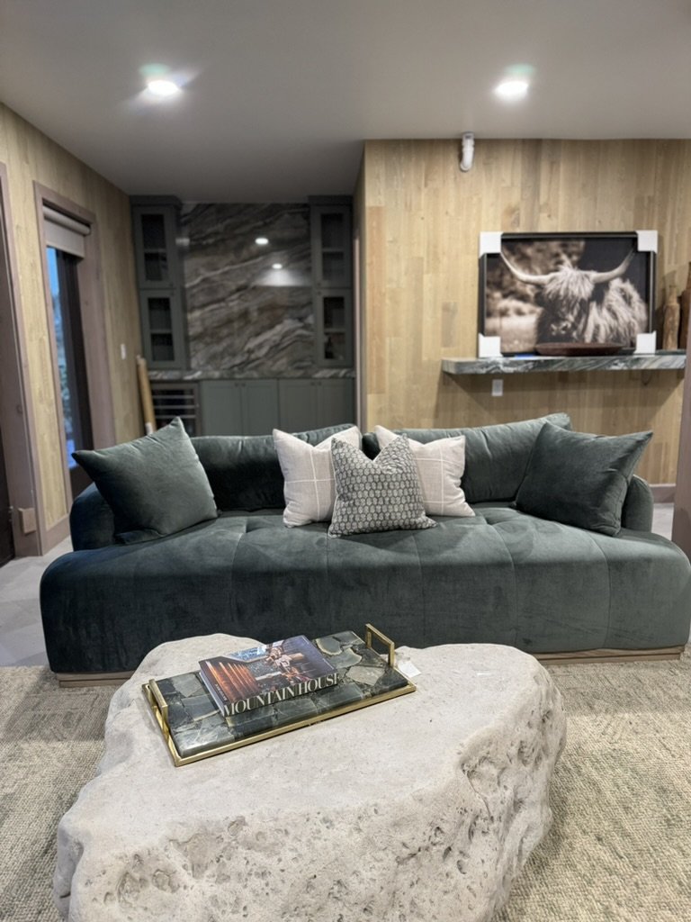Living room with a dark green sofa, decorative pillows, a stone coffee table with a tray and magazines, a large wall art of a highland cow, and a wooden wall with a marble shelf.