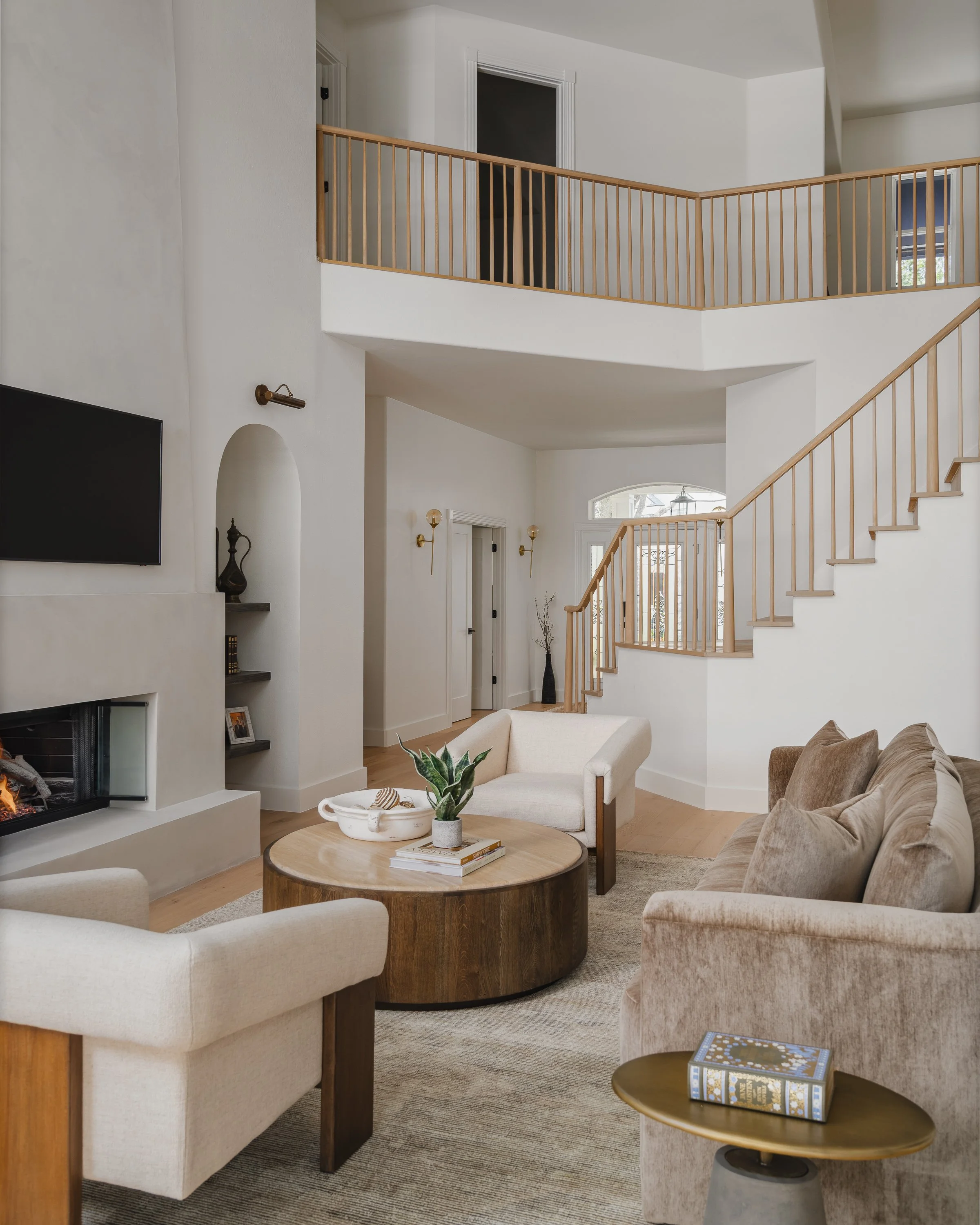 Living room with beige sofa, white armchair, wooden round coffee table with a plant, fireplace, flat-screen TV, and staircase leading to an upper level with wood railing.