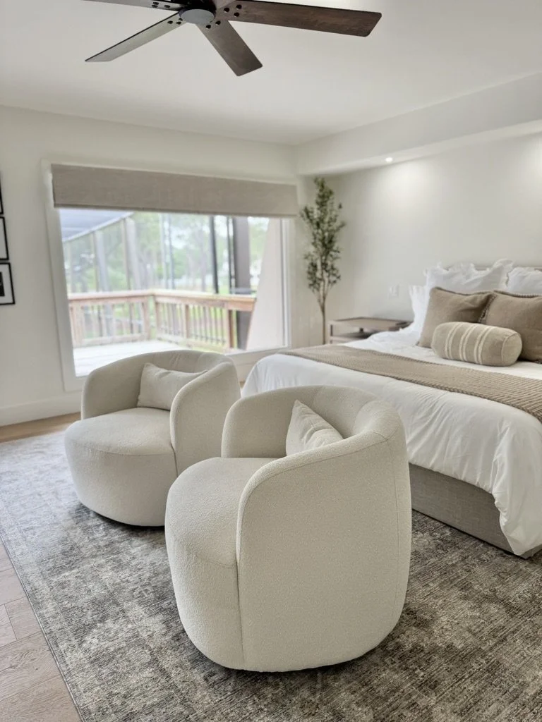 A bedroom with a large bed, two white upholstered armchairs with pillows, a boundary chain with an outdoor view, and a ceiling fan.