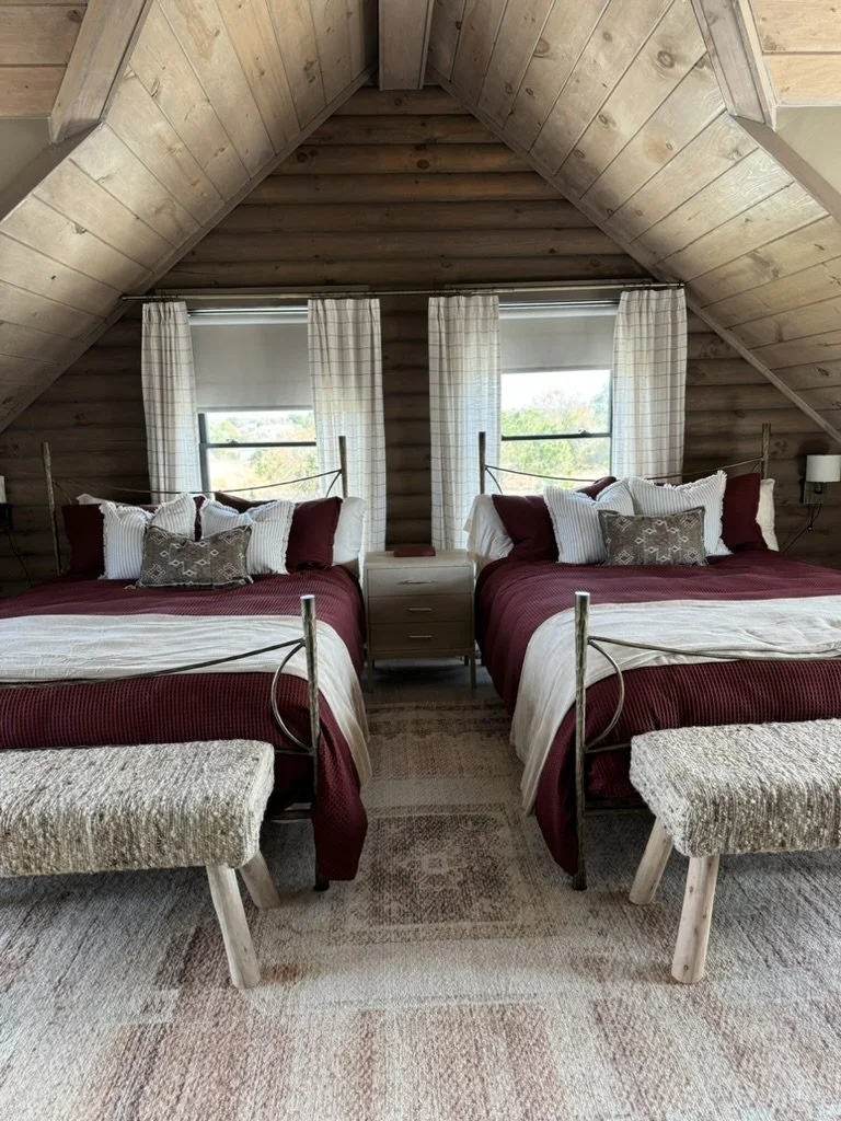Two beds with maroon and white bedding, pillows, and headboards, flanking a small nightstand, in an attic room with wood-paneled walls, two windows with curtains, and two matching benches at the foot of each bed.