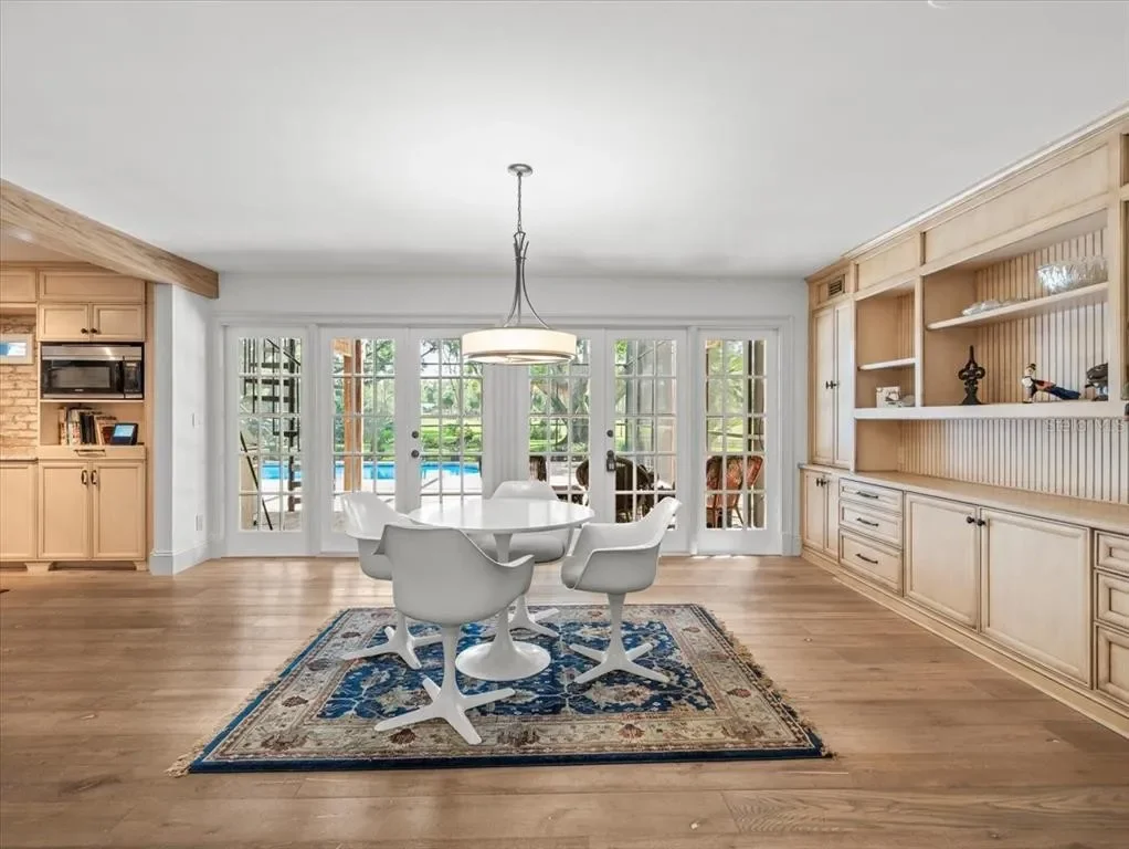 Bright dining room with a white round table, five white swivel chairs, a patterned rug, built-in wooden cabinets, and glass French doors leading to a backyard with a pool.