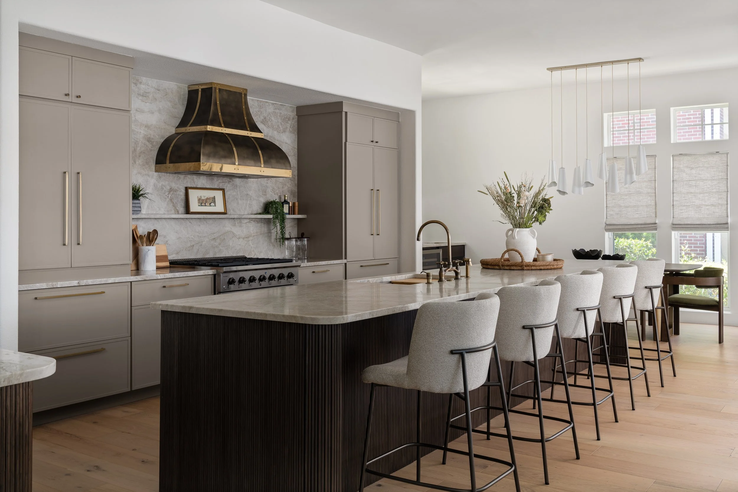 Modern kitchen with beige cabinetry, marble backsplash, black and gold range hood, large marble island with light gray chairs, flowers in a white vase, and large windows with Roman shades.
