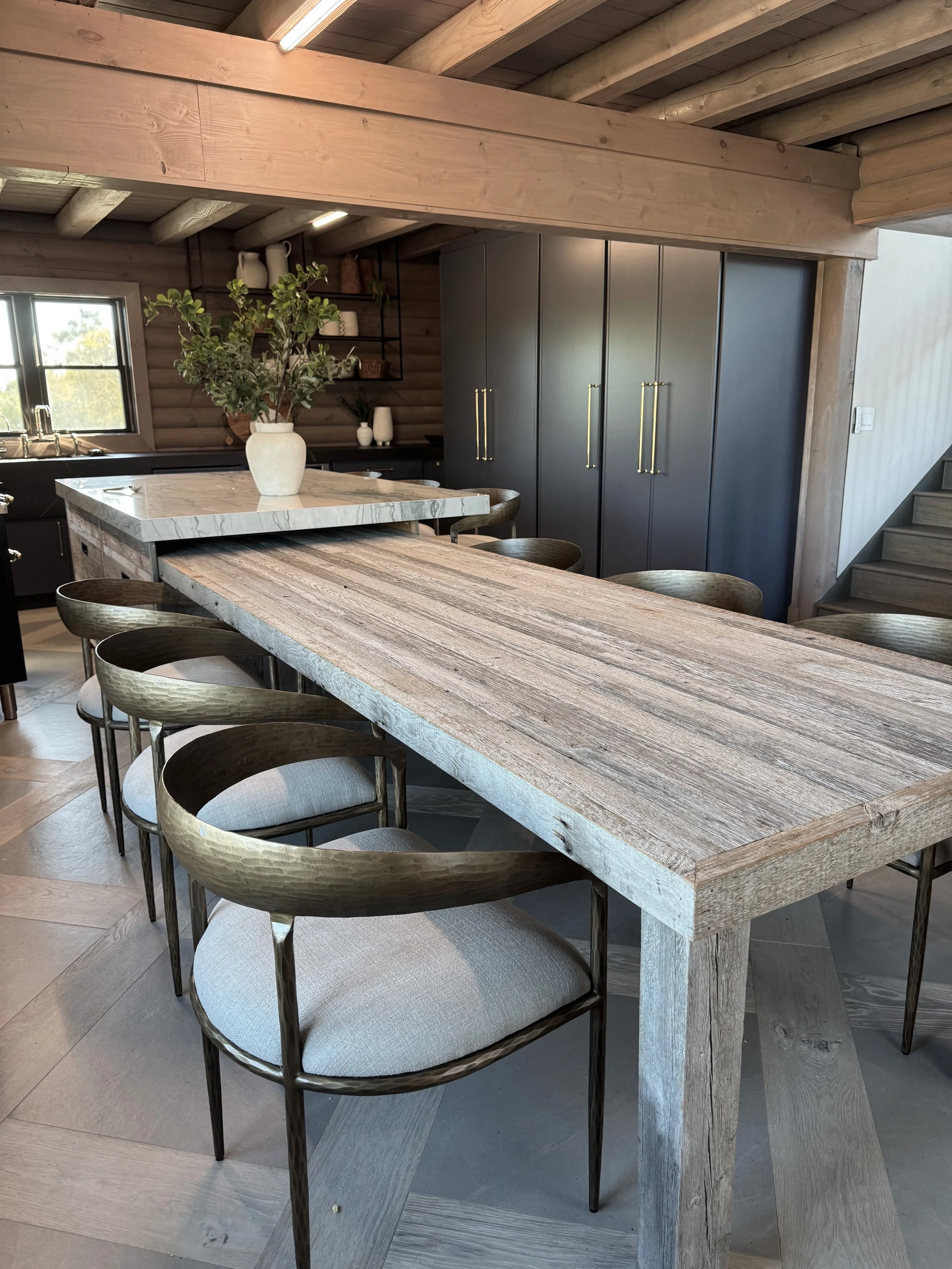 Modern kitchen with a large wooden dining table, six matching chairs with metal frames and cushioned seats, black cabinetry, open shelves with dishes, a window, and a large vase with greenery on the table.